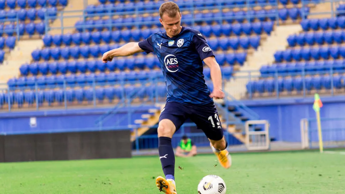 A soccer player on the field kicking a ball during a game or practice, with empty stadium seats in the background.