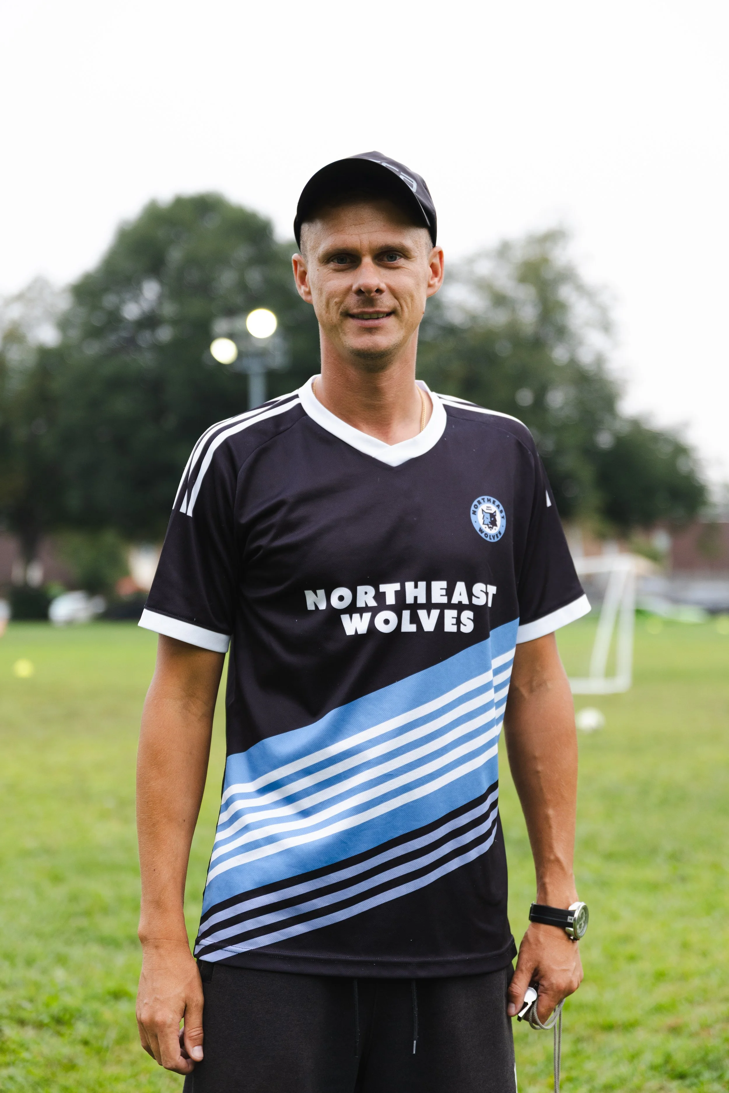 A man wearing a black and blue sports jersey with 'NORTHEAST WOLVES' written on it, standing outdoors on a grassy field with trees and cloudy sky in the background.