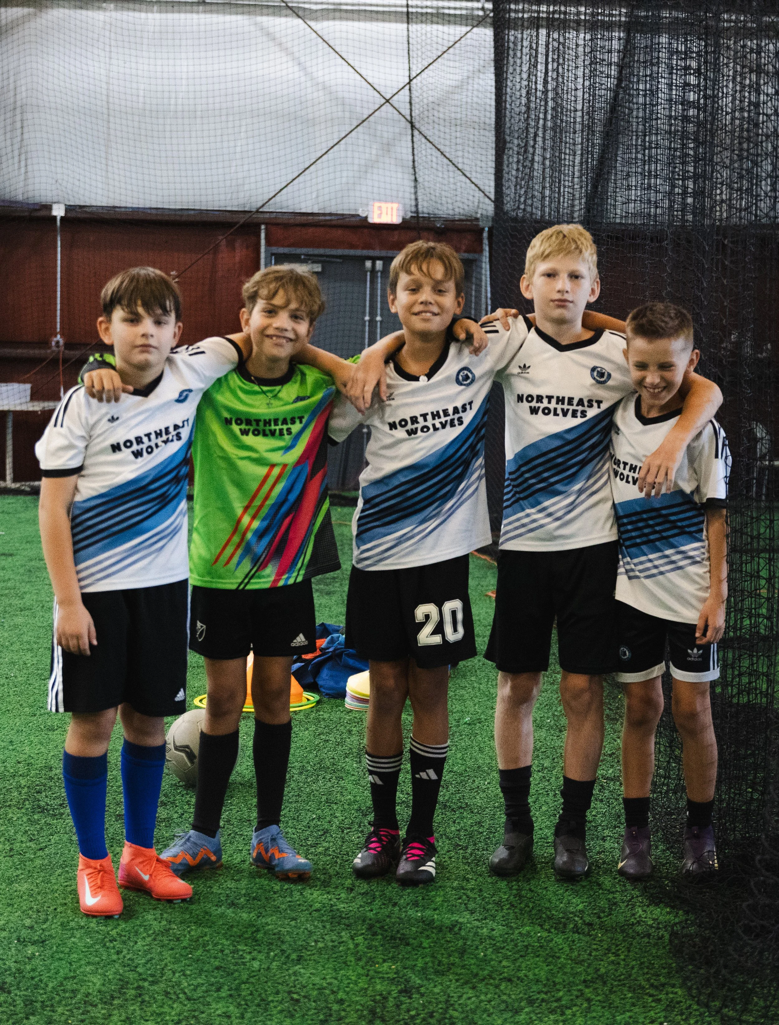 Five young boys standing together in soccer uniforms on a green turf indoor field, smiling with arms around each other.