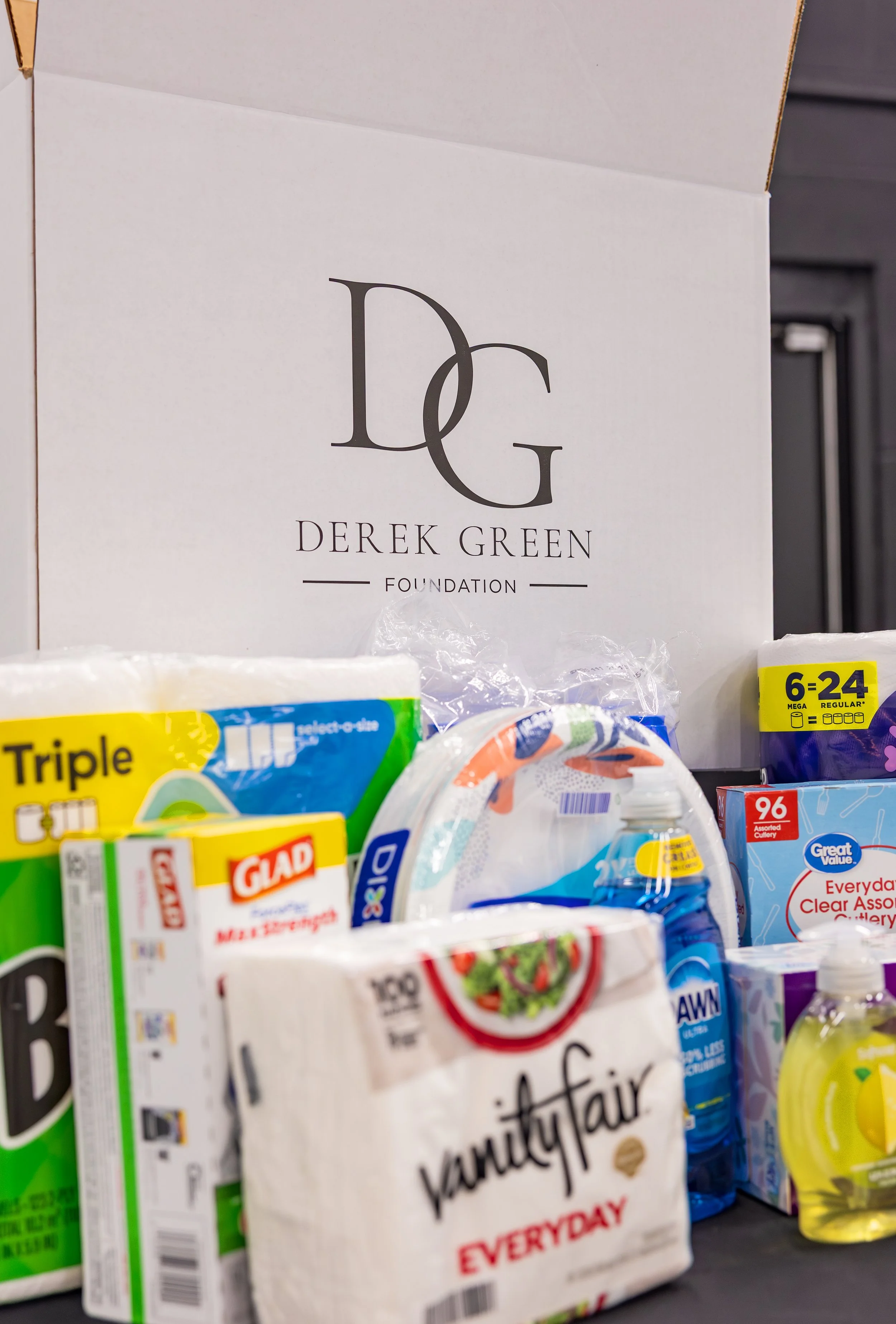 A white Derrick Green Foundation box on a shelf surrounded by household items including toilet paper, paper towels, dish soap, and other cleaning supplies.