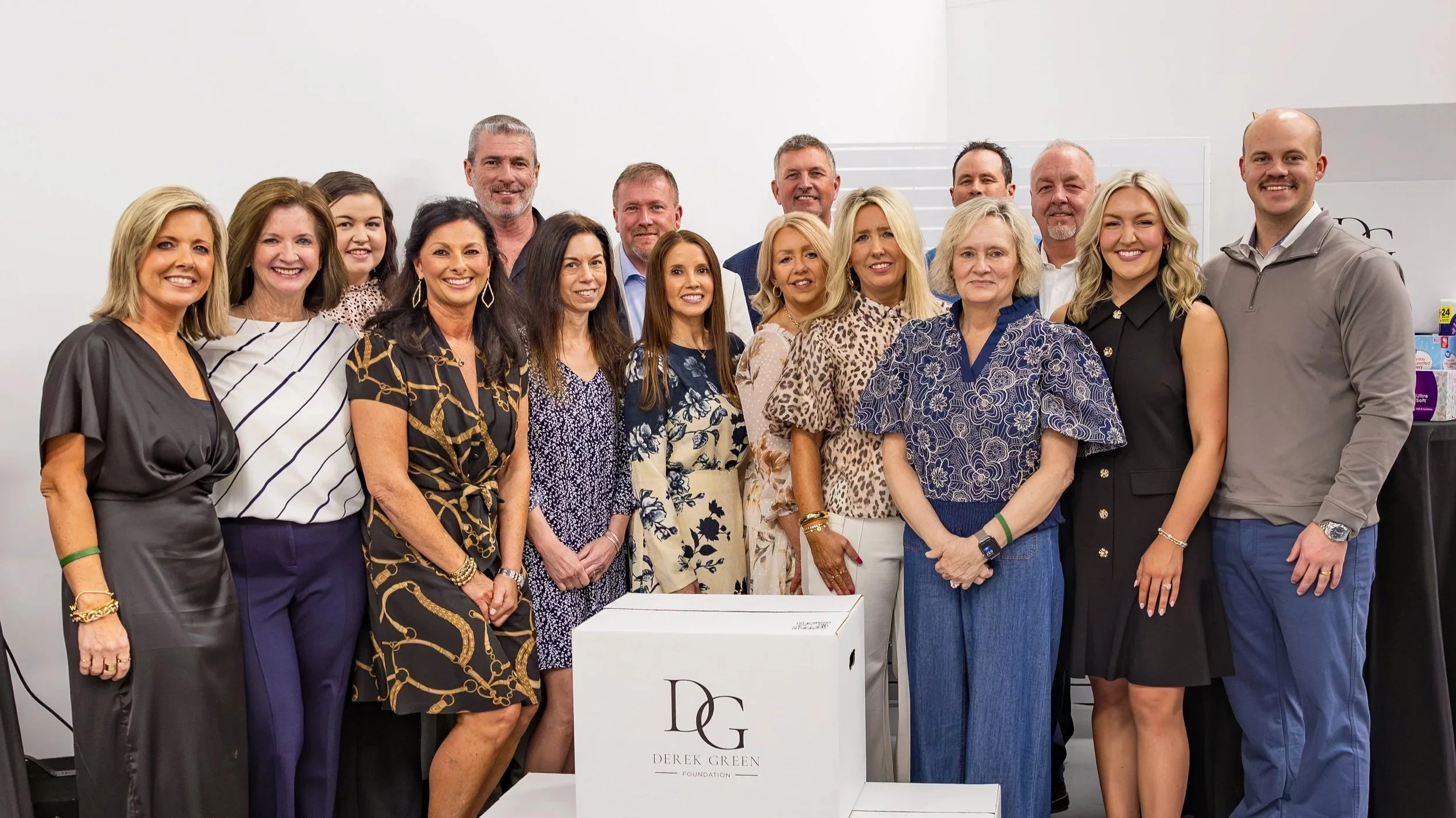 Group of professionally dressed people, men and women, standing together indoors at a charity or foundation event, with a white box labeled Derek Green Foundation in front of them.
