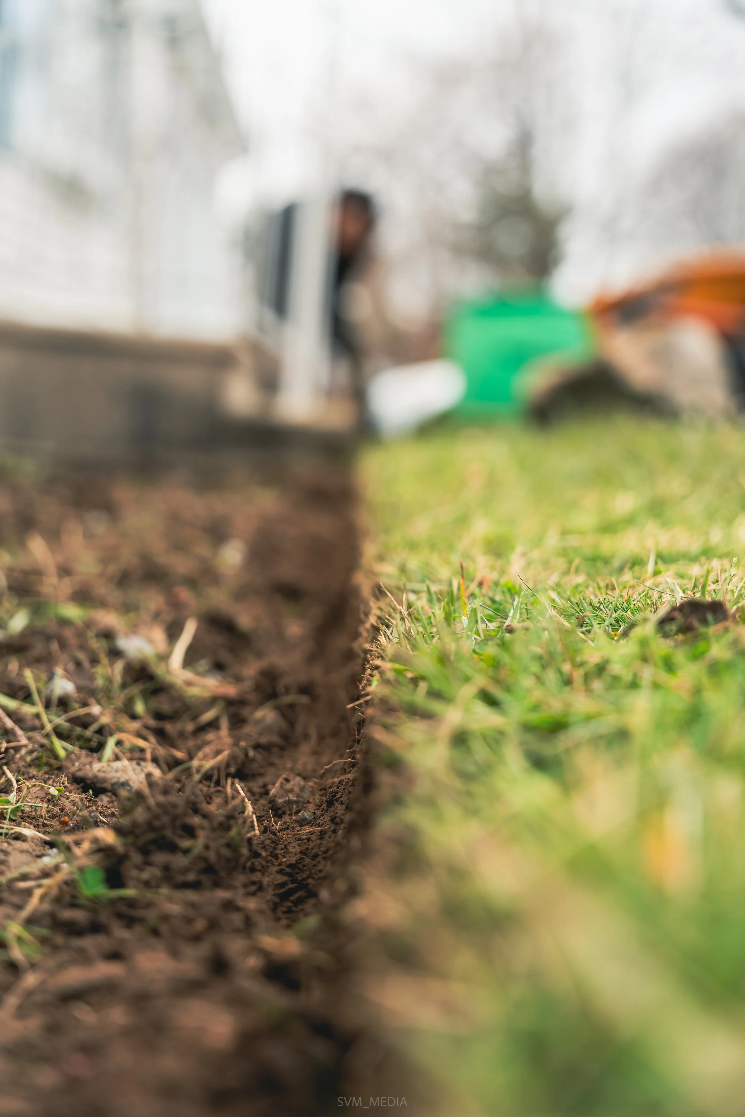 A close-up view of a freshly dug narrow trench in the ground with grassy surroundings, with a person operating construction equipment blurred in the background.