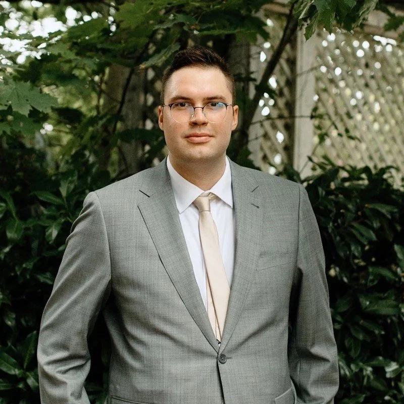 A young man in a gray suit and tie standing outdoors in front of greenery and a lattice fence.