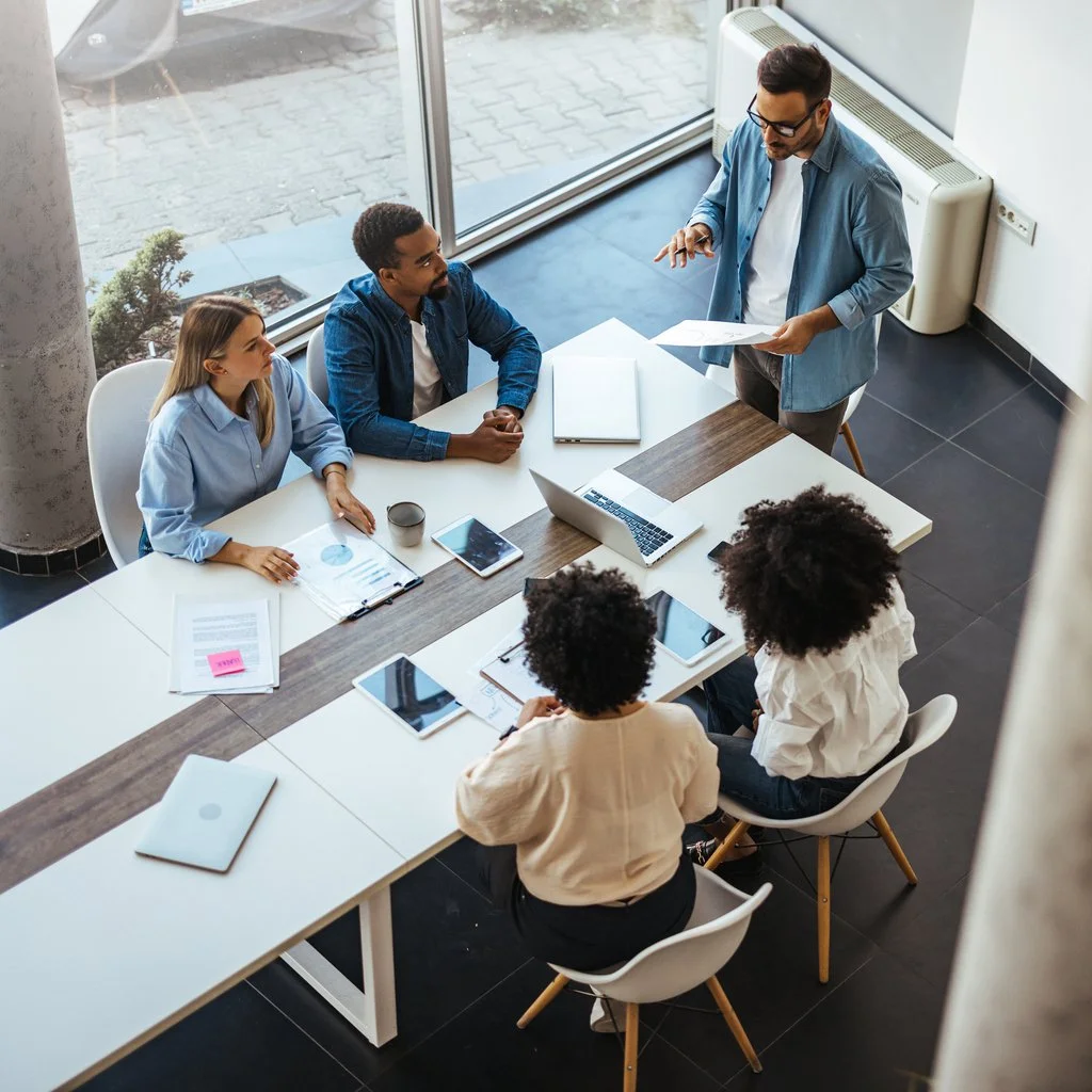 A group of five diverse people in a meeting room having a discussion around a rectangular table, with two women and three men, some taking notes or using electronic devices, while one standing man appears to be leading the conversation.