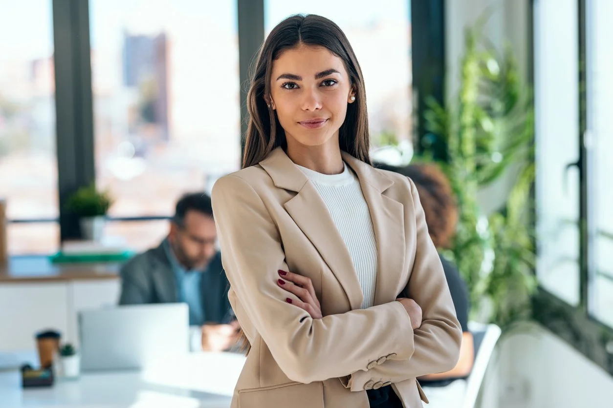 A young woman in a beige blazer standing confidently with arms crossed in a bright office space, with two colleagues working in the background.
