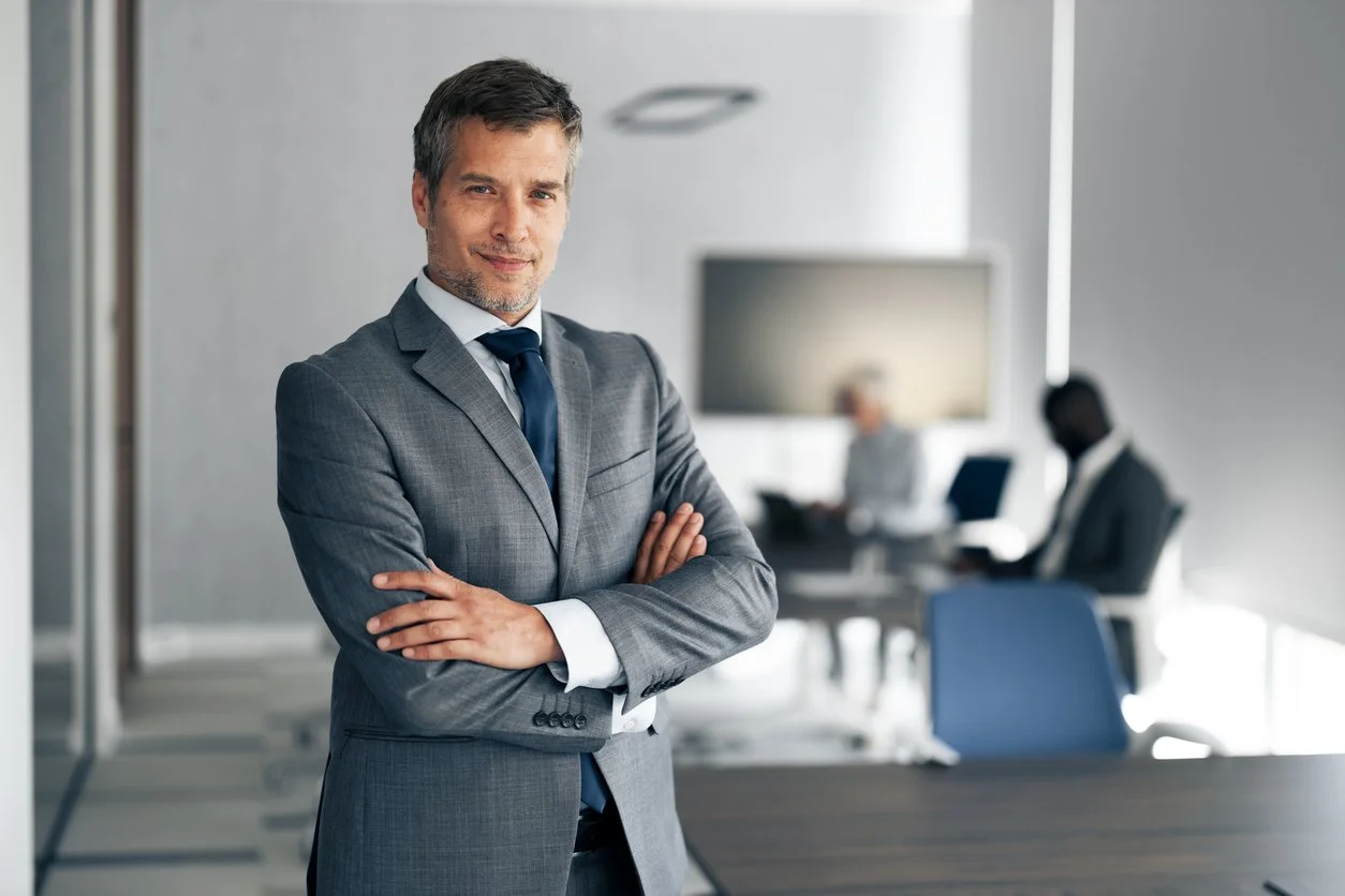A confident middle-aged man in a gray suit with a navy tie, arms crossed, standing in a modern conference room with two blurred colleagues in the background.