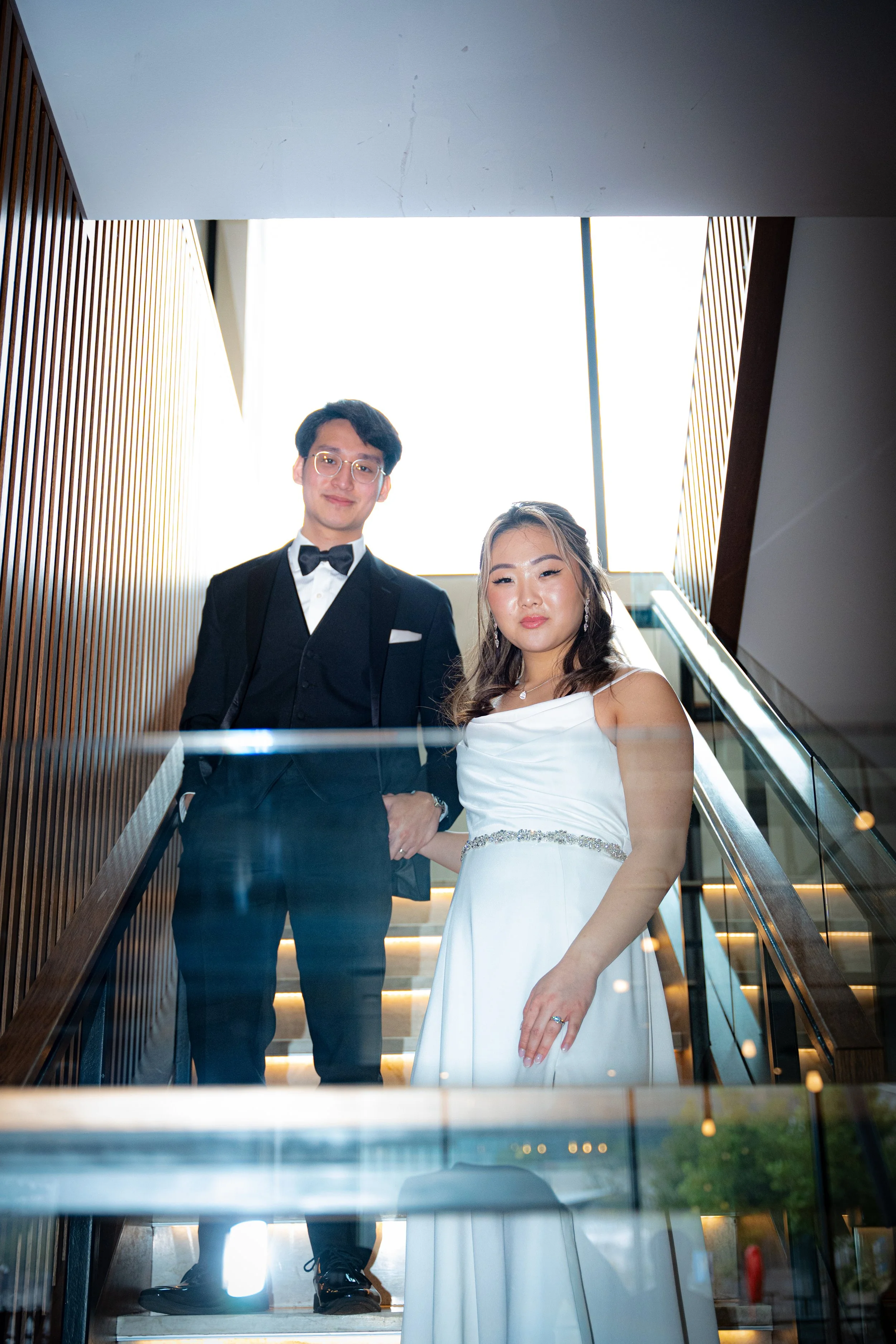A young couple dressed in formal attire standing on a staircase, with the man in a black tuxedo and the woman in a white gown, holding hands and posing for a photo.