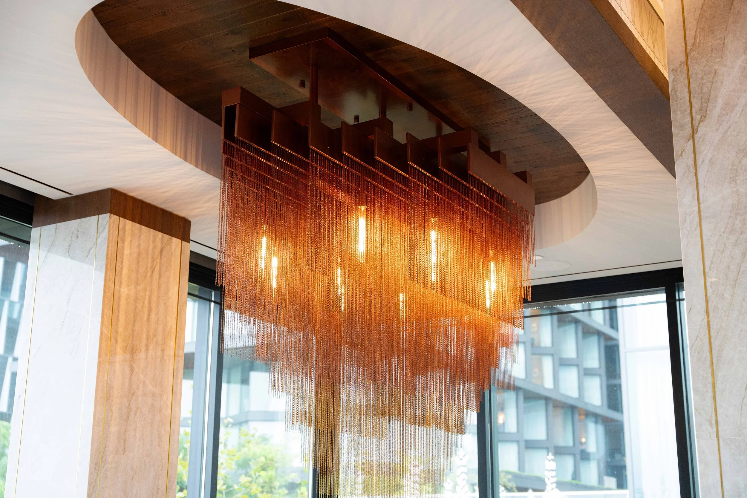 A modern chandelier with numerous hanging metal chains and vertical light bulbs inside a building with large windows and marble walls.