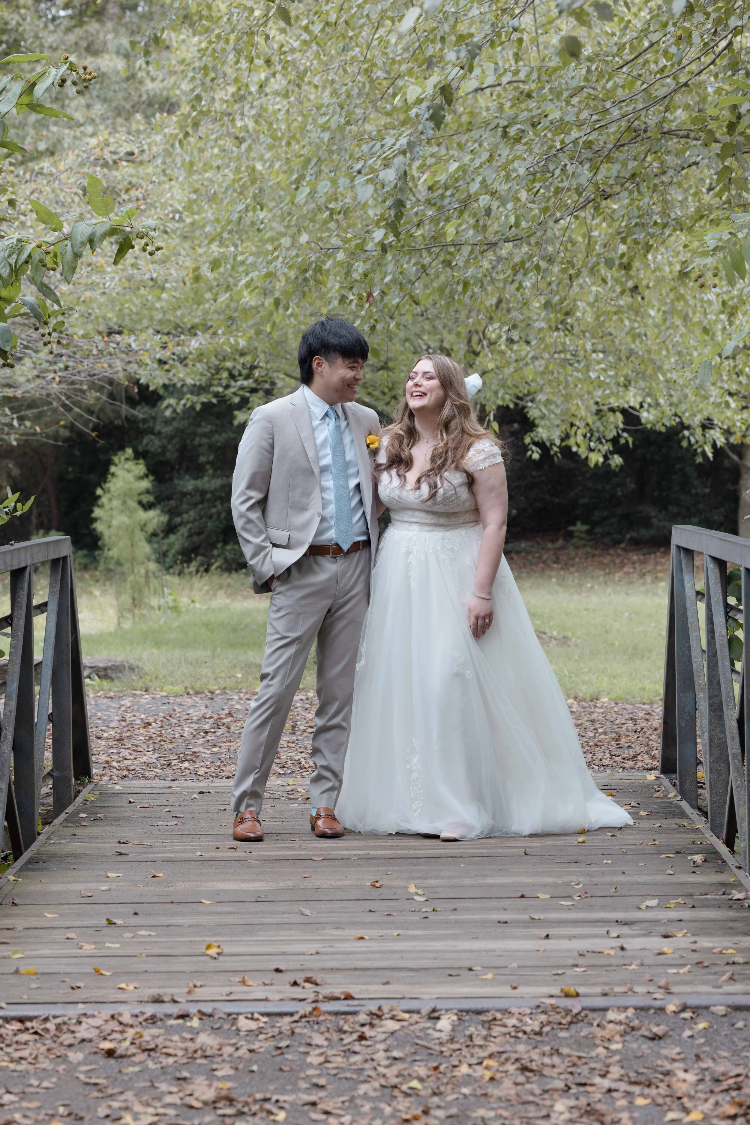 A bride and groom standing on a wooden bridge in a park, smiling and looking at each other surrounded by green trees and fallen leaves.