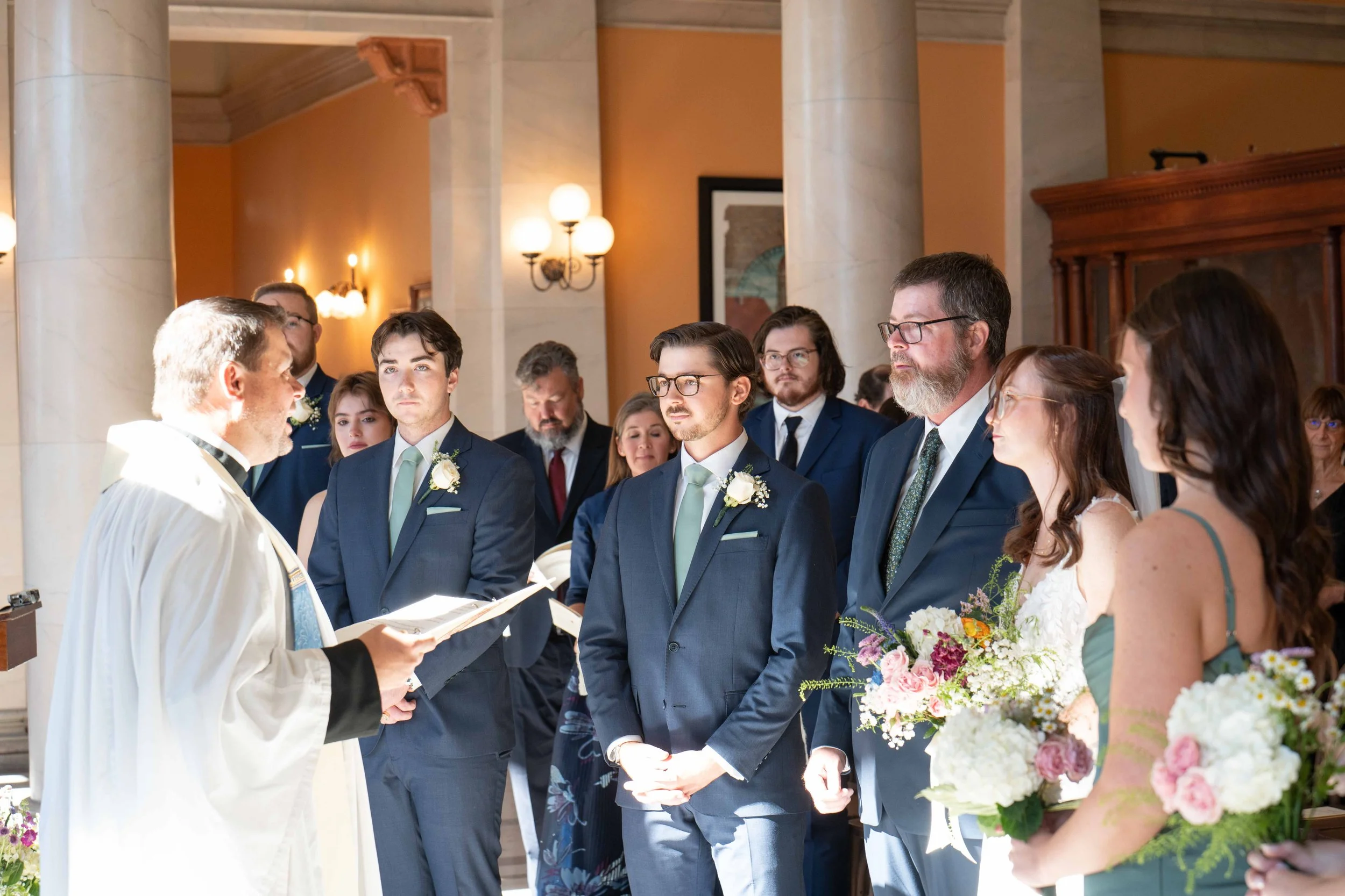 A wedding ceremony with a priest officiating, standing in front of the bride and groom and their wedding party, inside a church with warm lighting and marble columns.