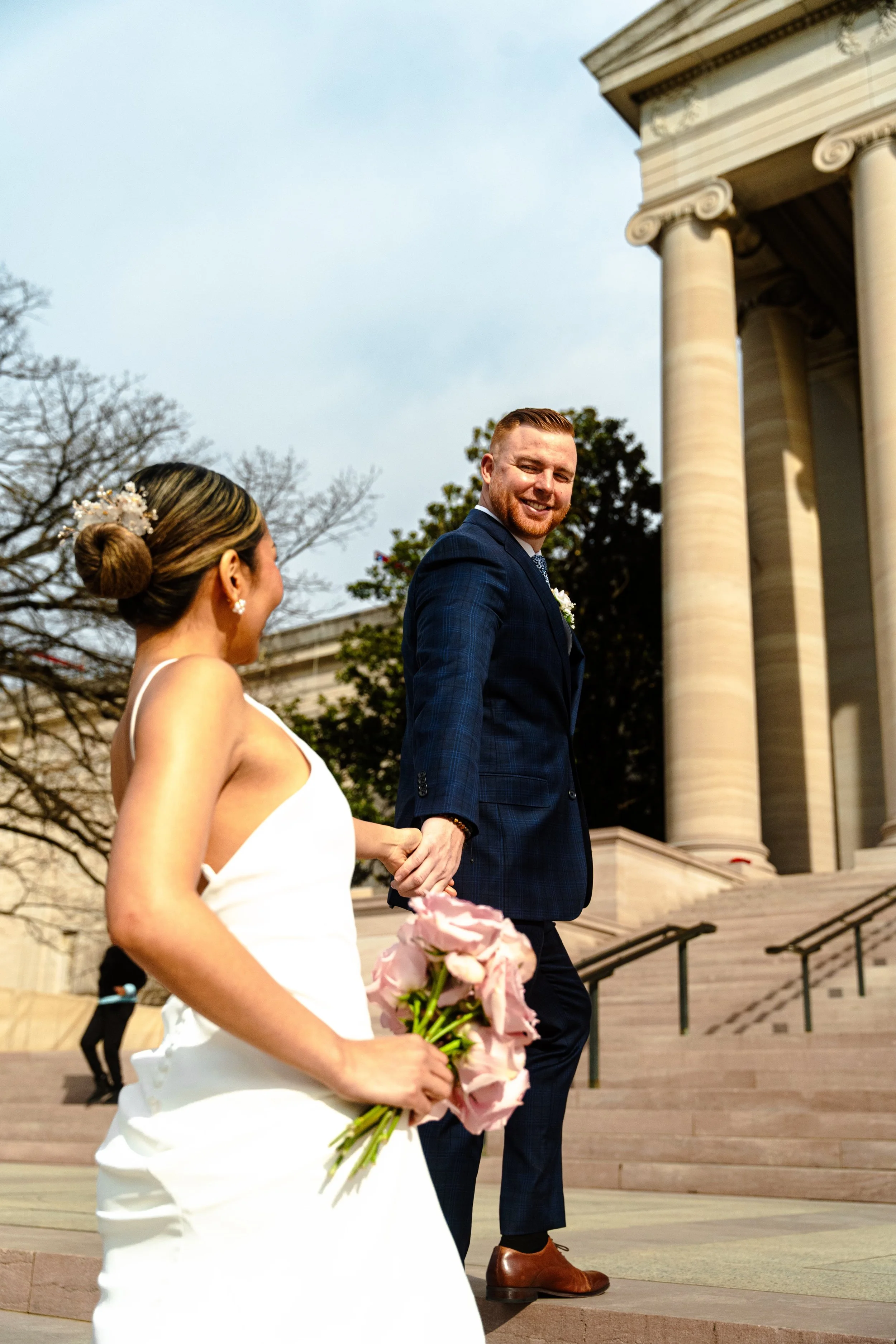 A bride in a white wedding gown holding a pink bouquet walking hand-in-hand with a groom in a dark blue suit at a wedding ceremony outdoors near a historic building with large columns.