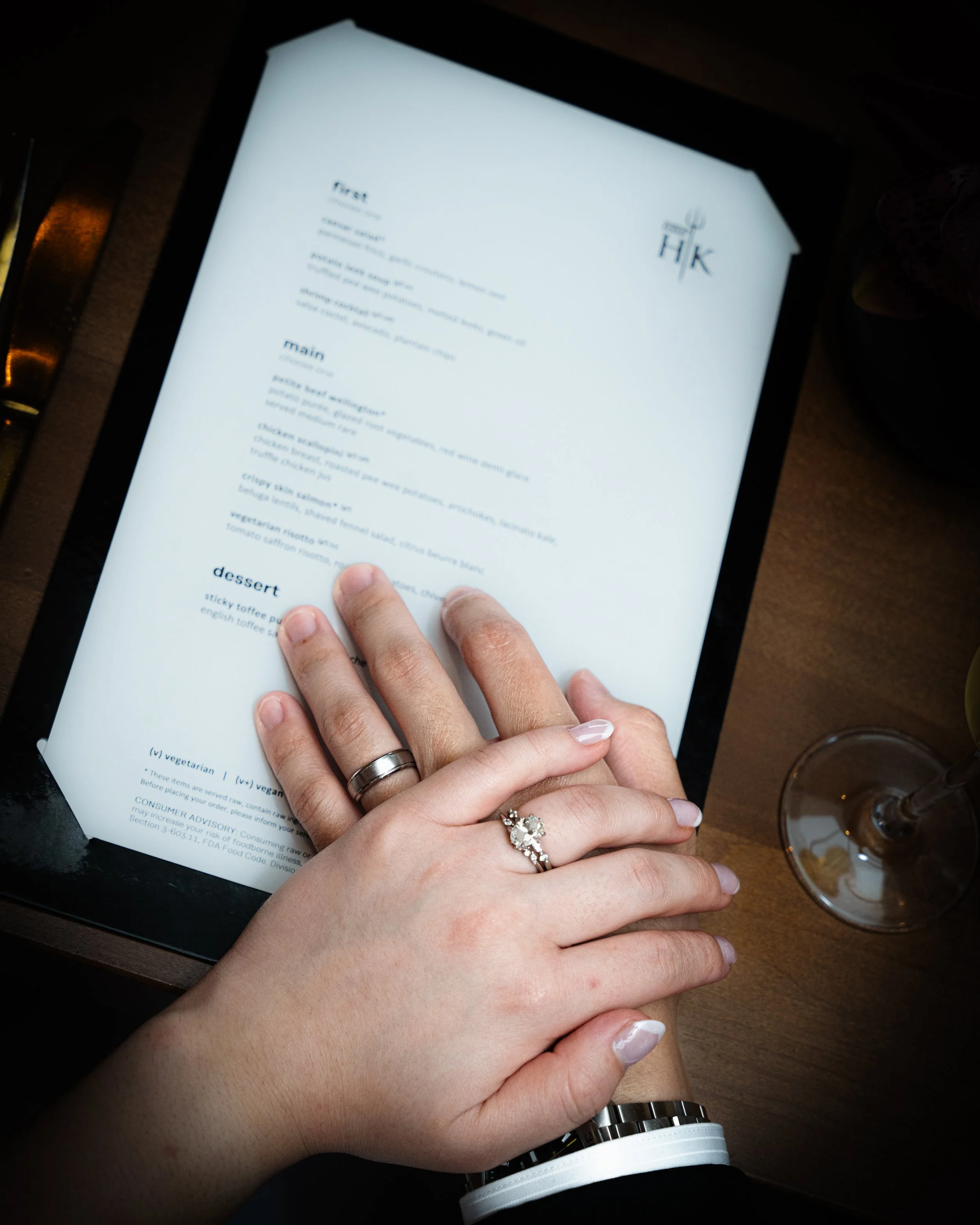 Close-up of a person's hand resting on top of another person's hand, both wearing rings, with a menu on a table in the background.