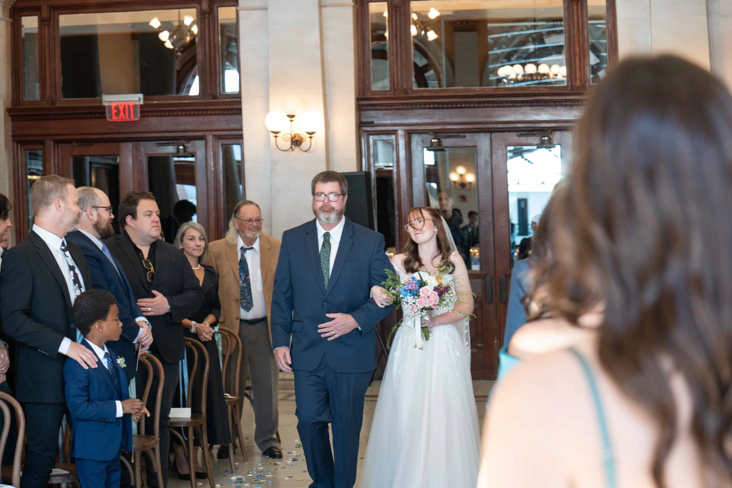 Bride walking down aisle with her father at a wedding ceremony, surrounded by guests sitting and standing in a decorated indoor venue.