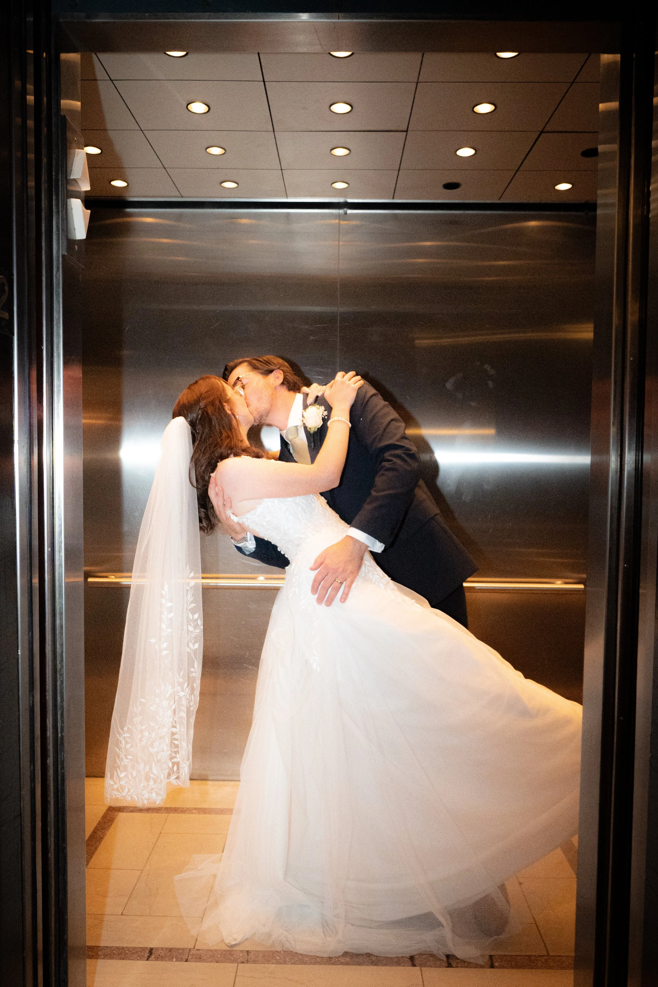 A newlywed couple sharing a kiss inside an elevator; the groom is dipping the bride who wears a white wedding gown and veil, and the groom is dressed in a black suit.