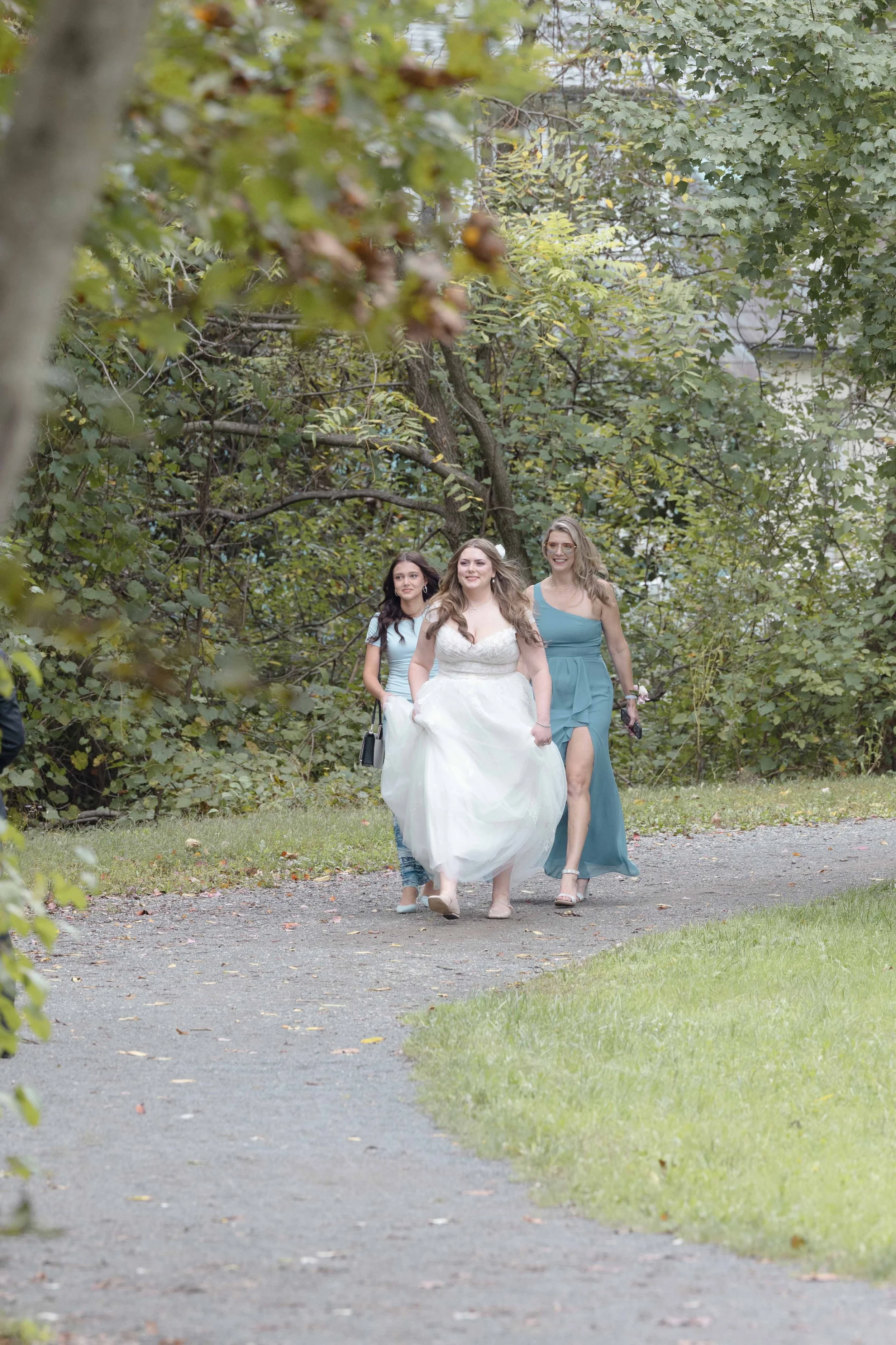 A bride in a white wedding dress walking on a park trail with two women, one in a blue dress and one in casual attire, surrounded by green trees.
