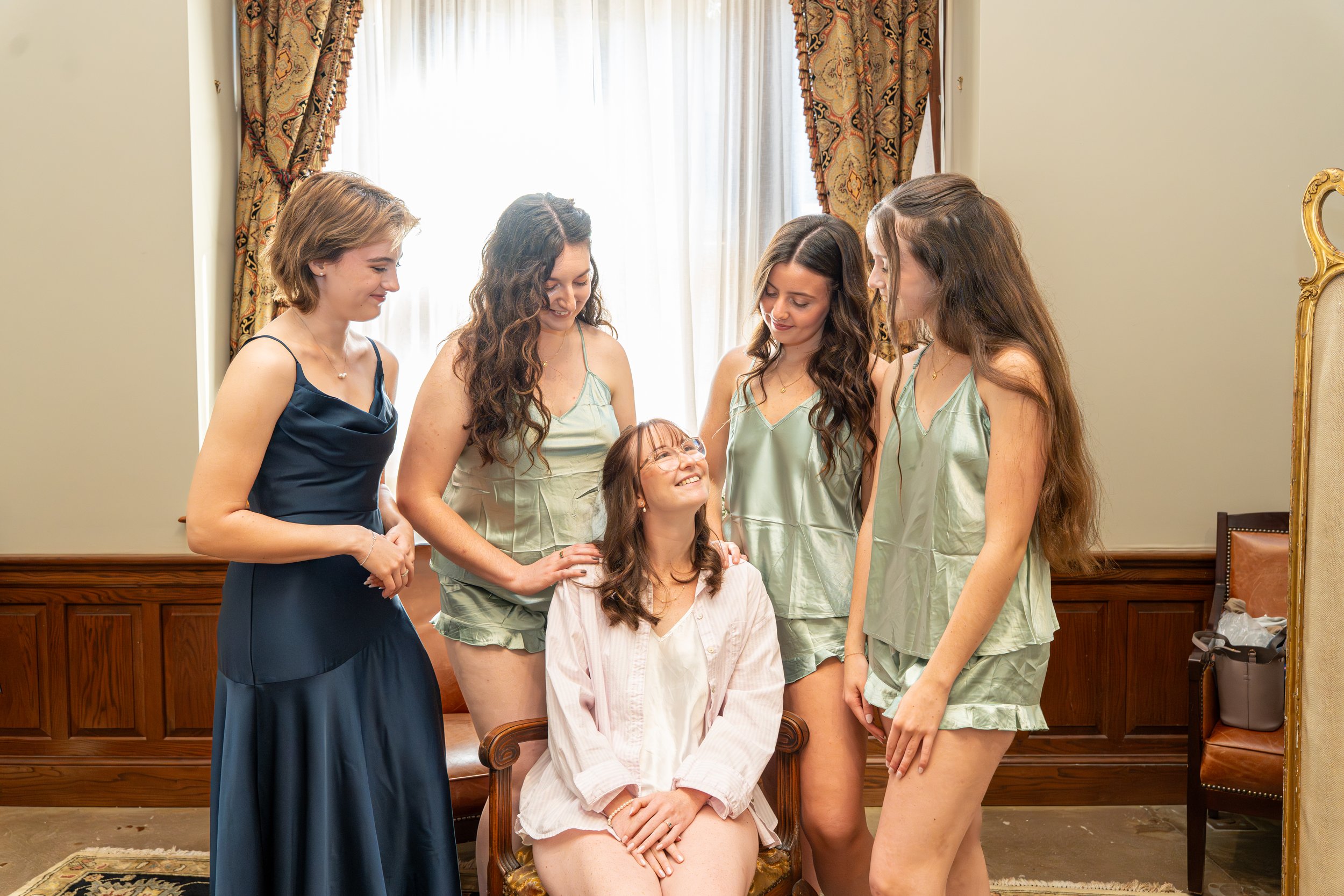 Six women, one seated and five standing, smiling and looking at each other in a warmly lit room with wooden paneling and decorated curtains.