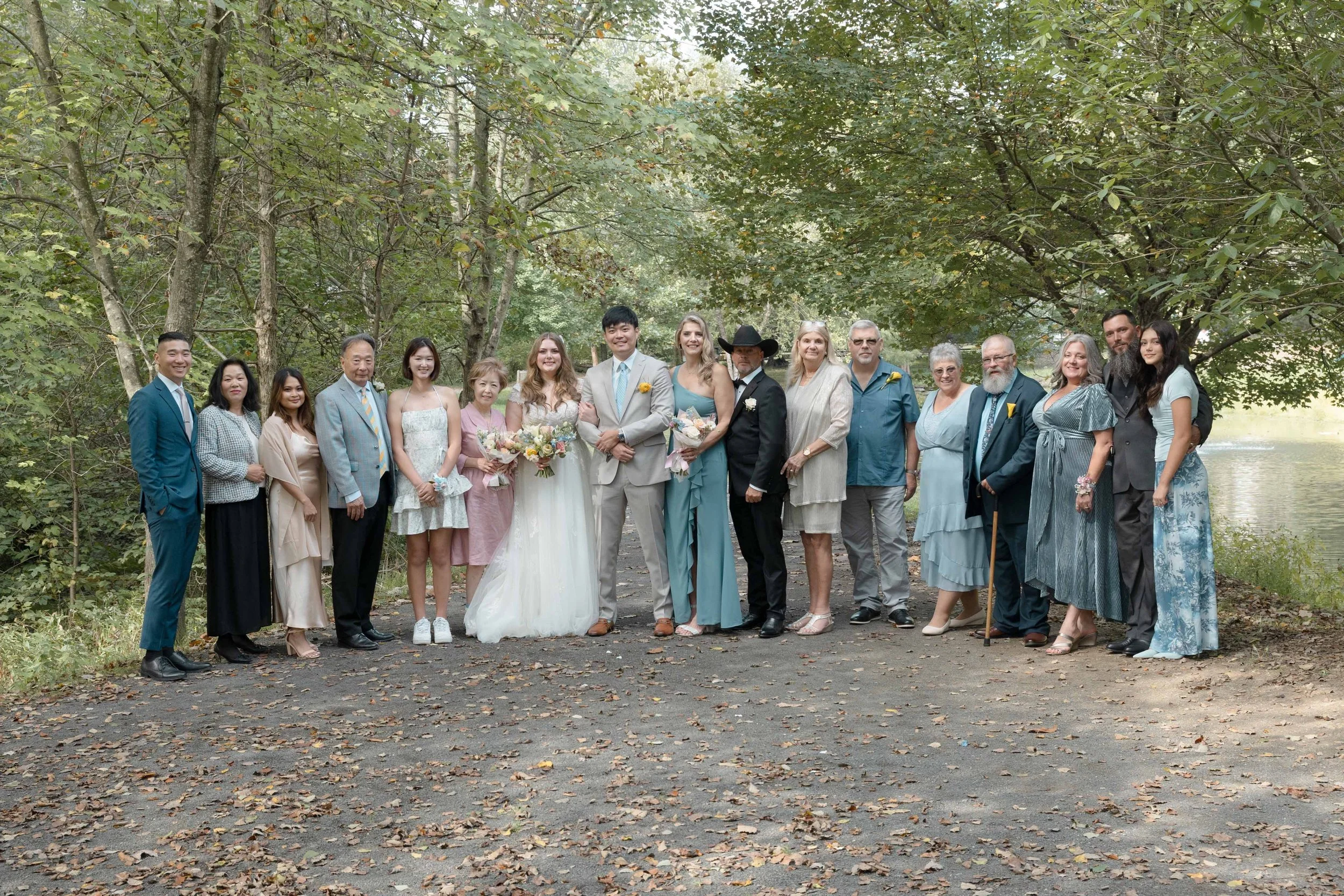 A group of people, including bride and groom, standing outdoors on a wooded path near a body of water, dressed in wedding attire and casual clothing, celebrating a wedding.