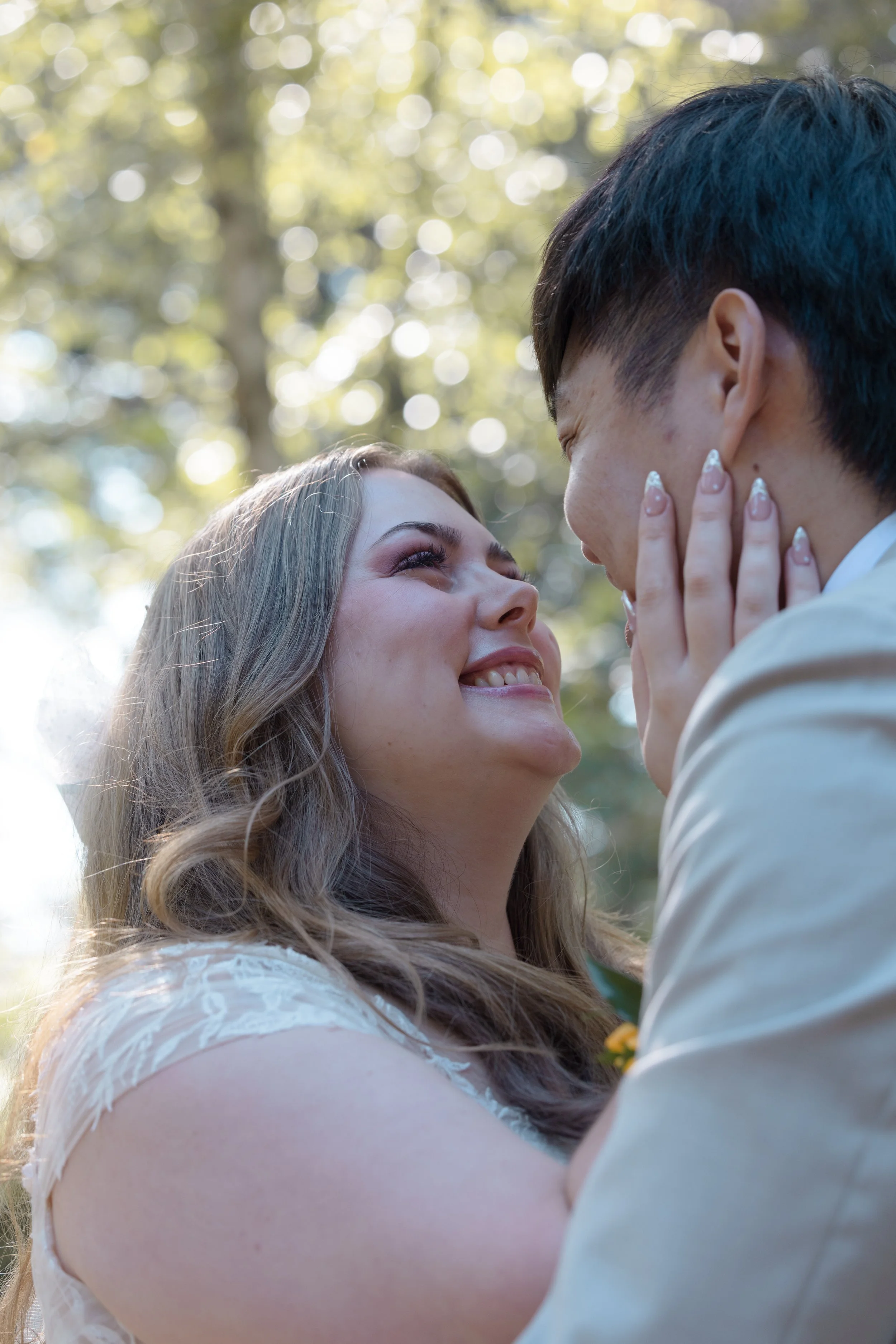 A smiling woman and man looking into each other's eyes outdoors with sunlight and trees in the background.