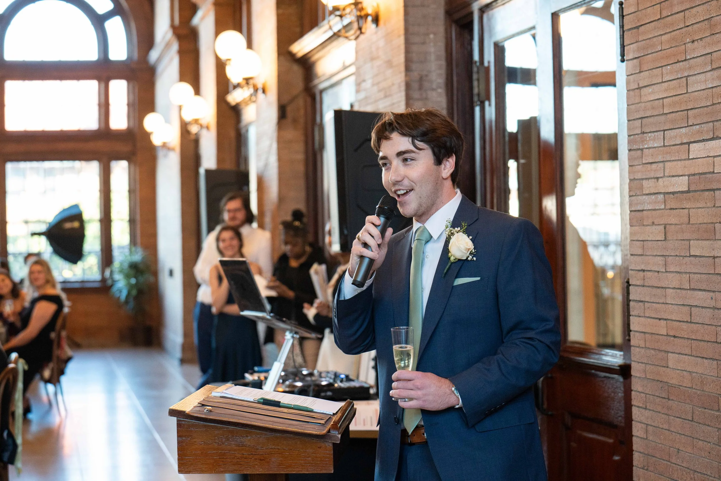 A man in a blue suit with a white shirt and a light green tie, holding a glass of champagne and speaking into a microphone at a wedding reception. He is standing next to a wooden podium with papers and a pen, with guests and a DJ in the background inside a warmly lit, brick-walled venue.