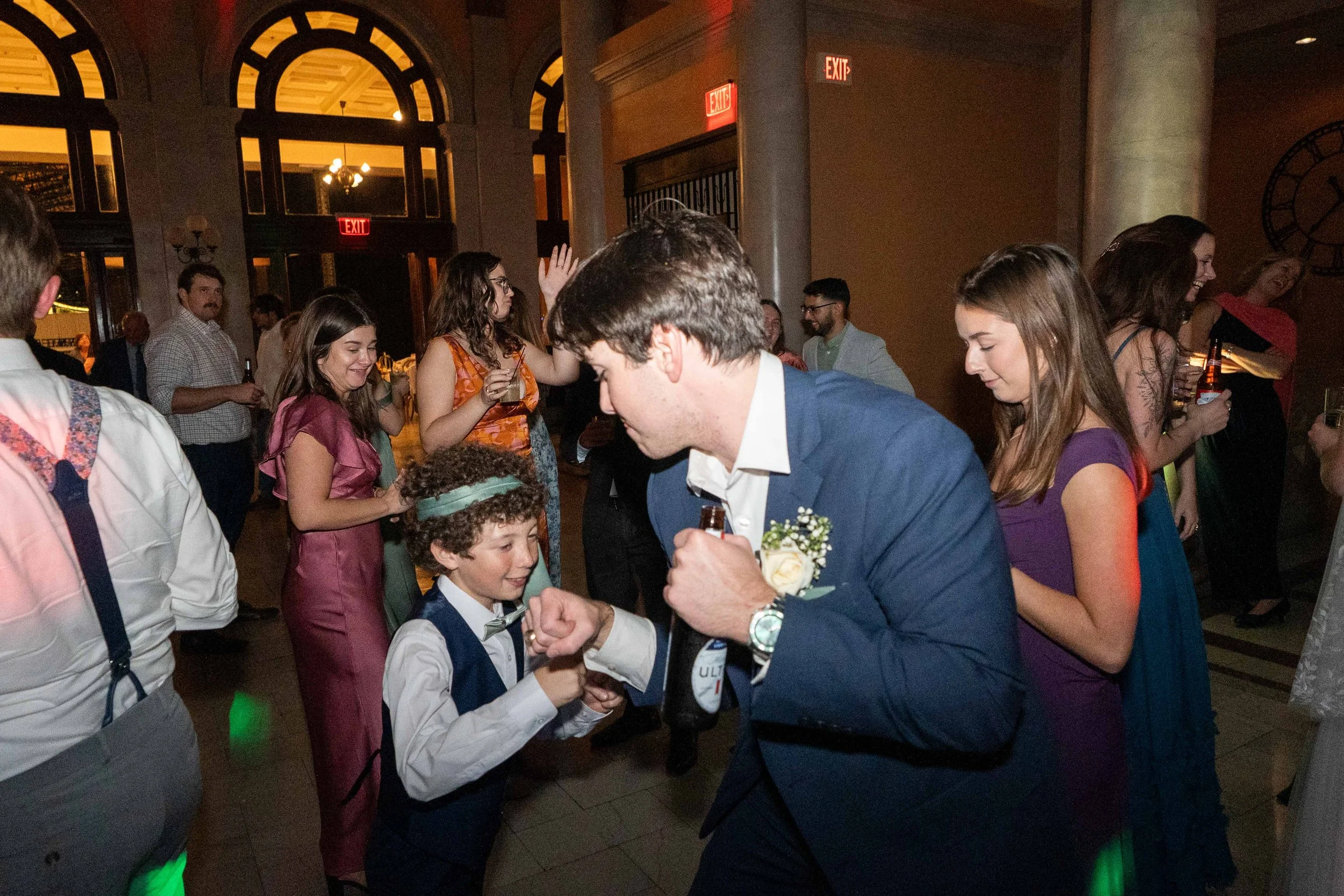 People dancing and socializing at a social event or party in a spacious, ornate room with large arched windows and warm lighting.