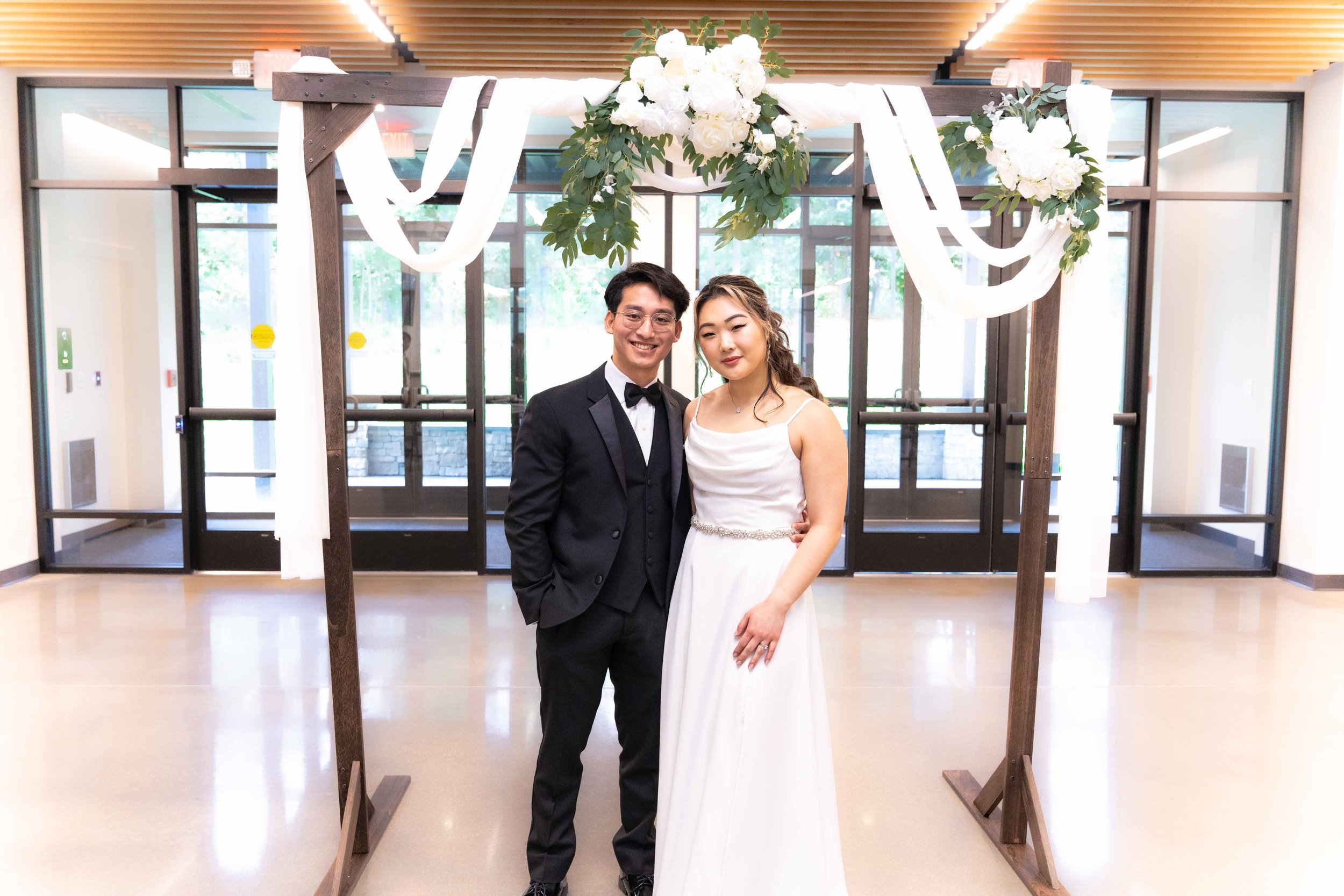 A bride and groom standing under a floral wedding arch, smiling at the camera, indoors with glass doors in the background.