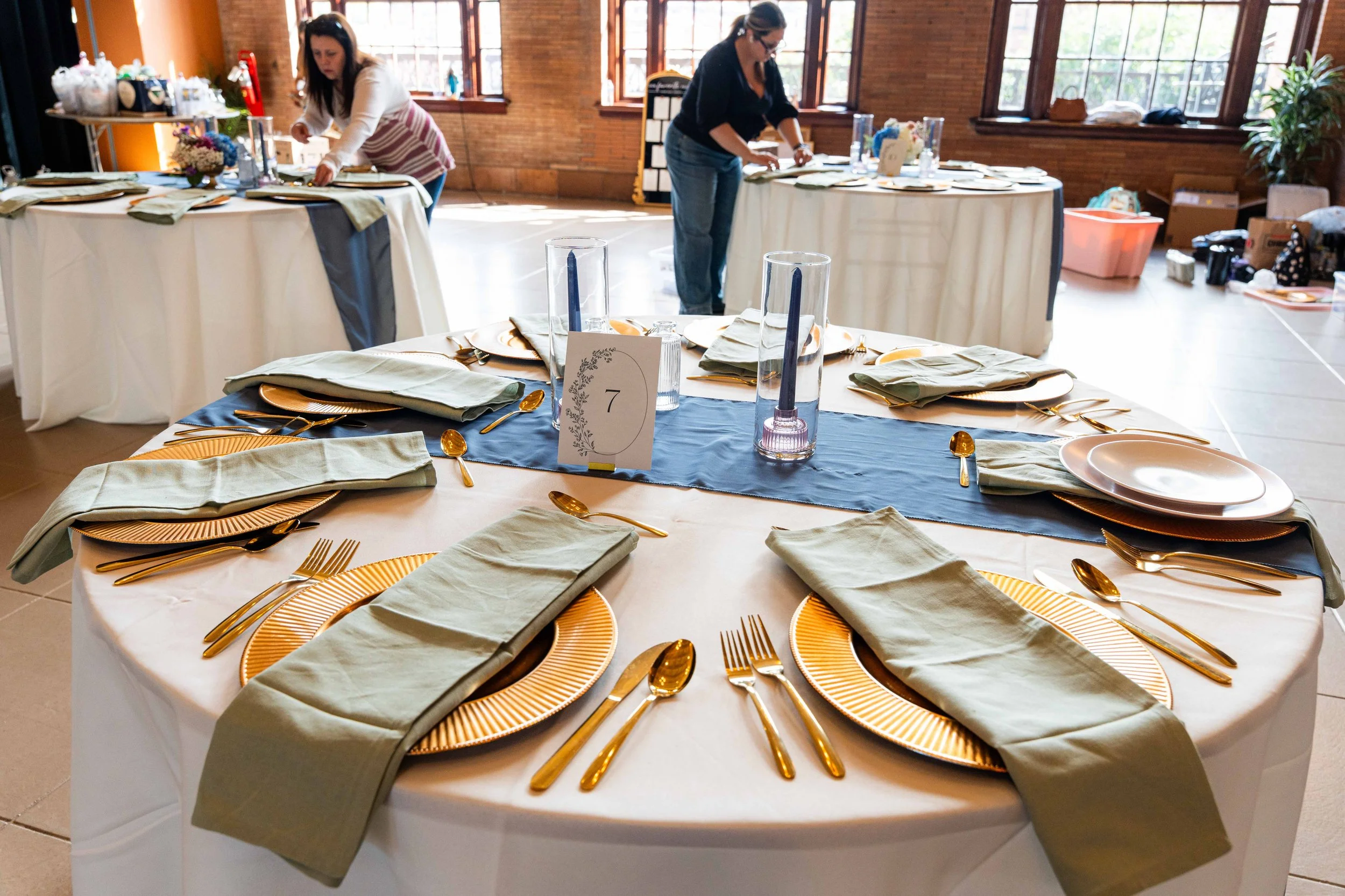 Round banquet table set for a formal event with gold utensils, beige napkins, orange plates, a dark blue table runner, a table number card, and tall glass candle holders, in a bright room with large windows and two women preparing additional tables in the background.