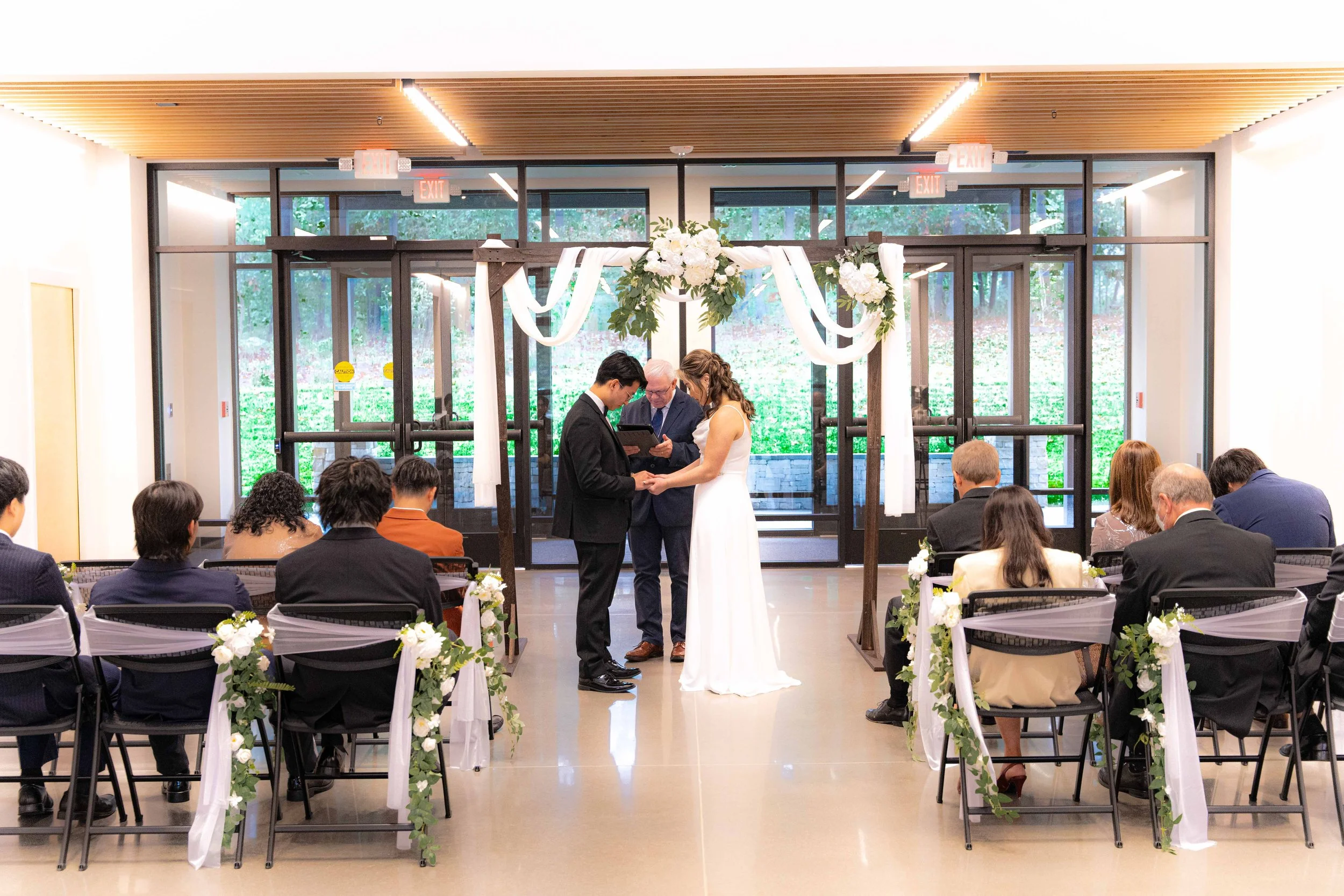 A wedding ceremony indoors with a bride and groom exchanging vows in front of an officiant, surrounded by seated guests, decorated with white flowers and drapery.