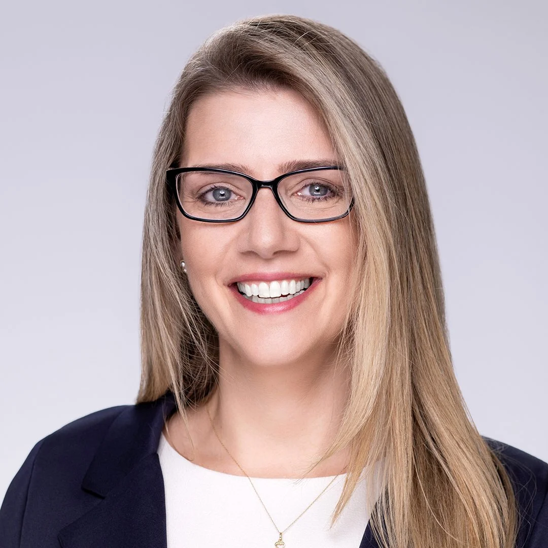 A smiling woman with blonde hair and glasses, wearing a dark blazer and a white top, posing against a plain background.