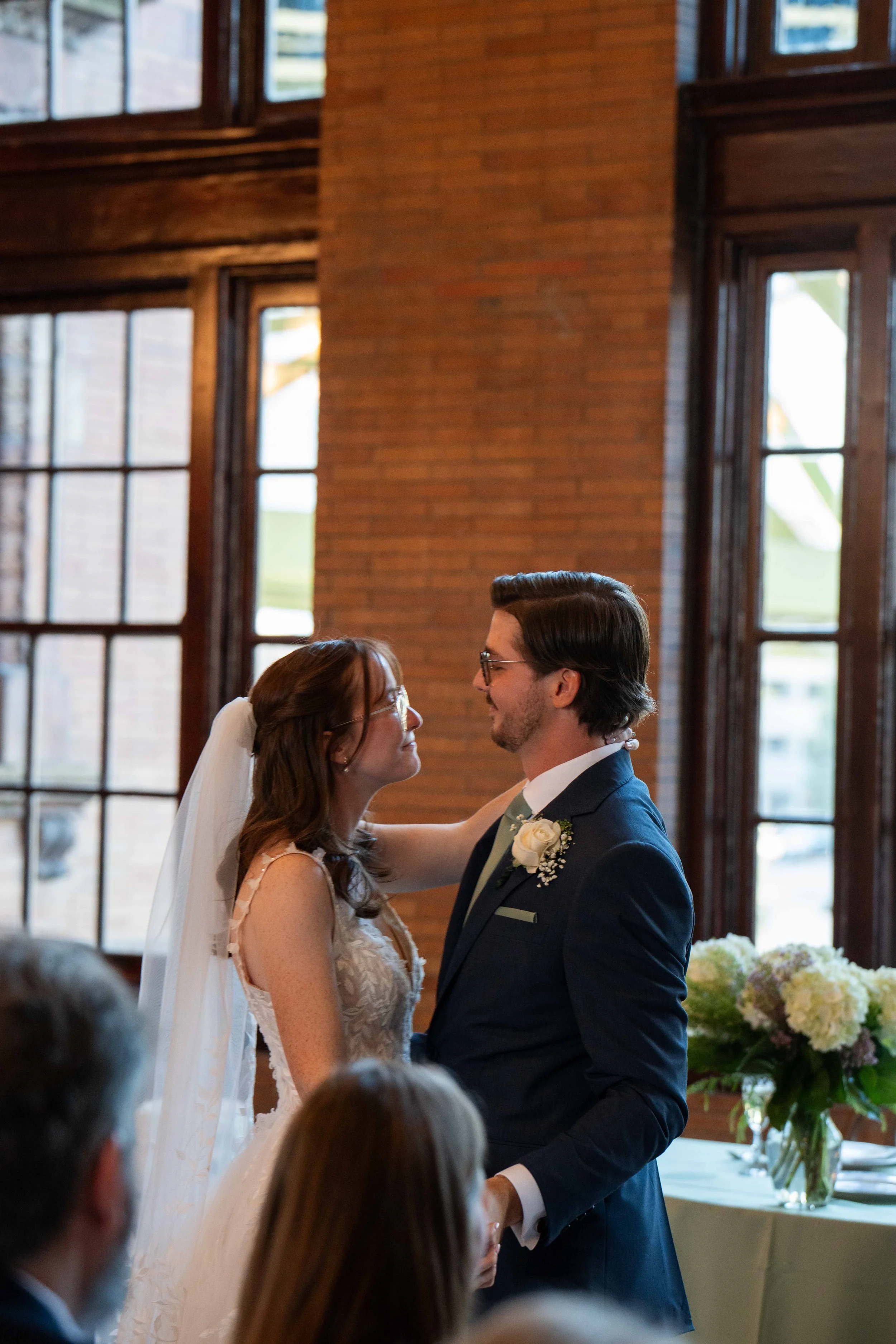 A bride and groom standing close, gazing into each other's eyes, during their wedding ceremony inside a venue with large windows and brick walls.