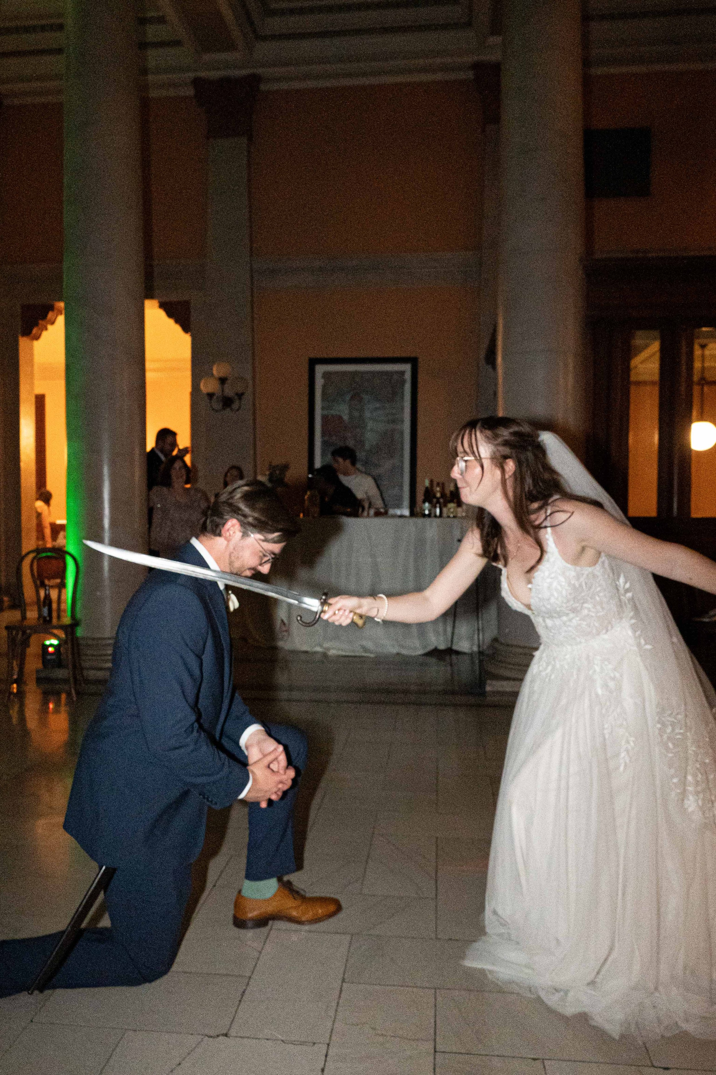 A bride and groom engage in a humorous sword fight during their wedding reception, with the bride playfully holding a sword to the groom's neck.