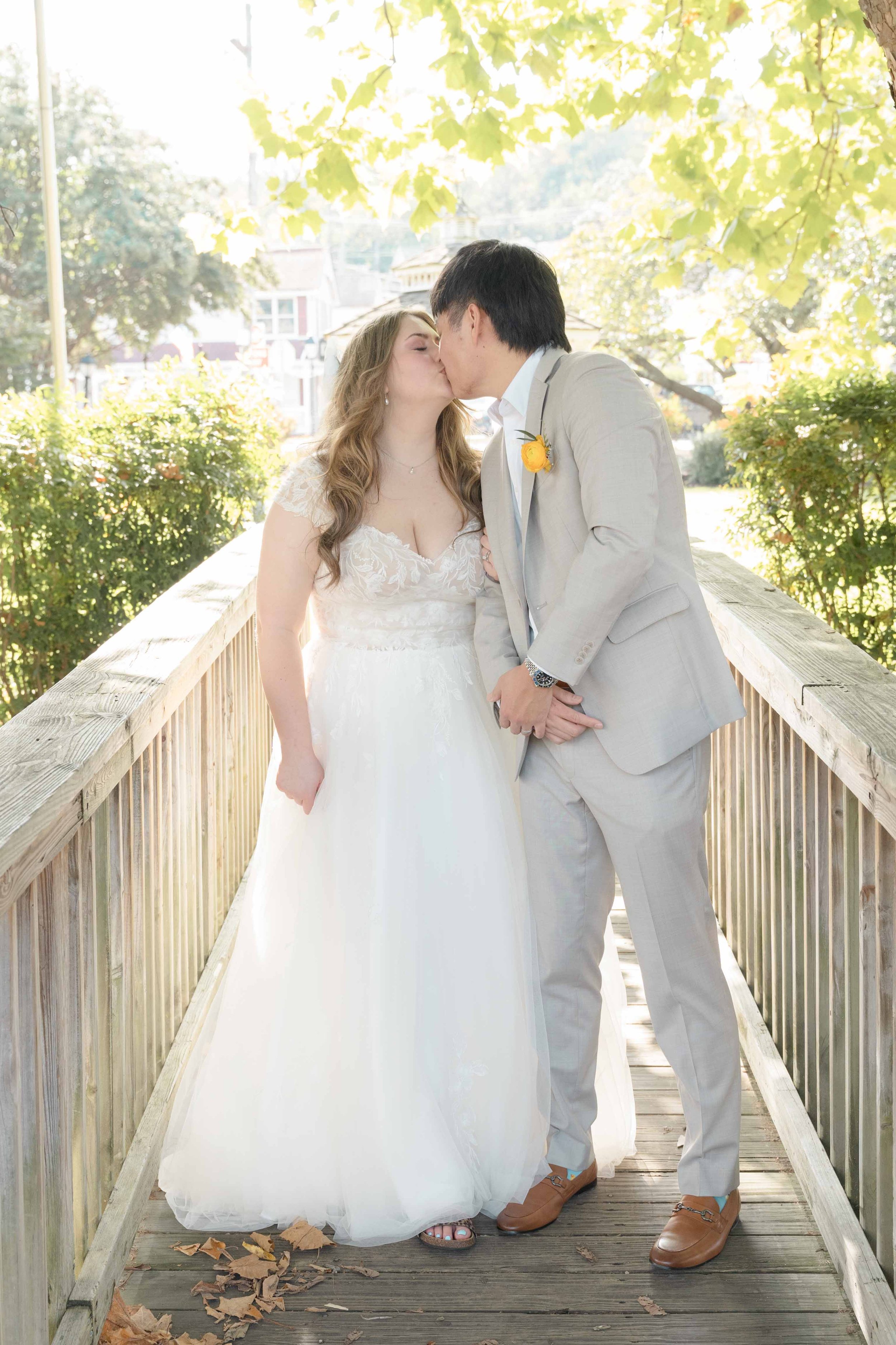 A bride and groom sharing a kiss on a wooden bridge outdoors, surrounded by greenery and sunlight, with houses in the background.