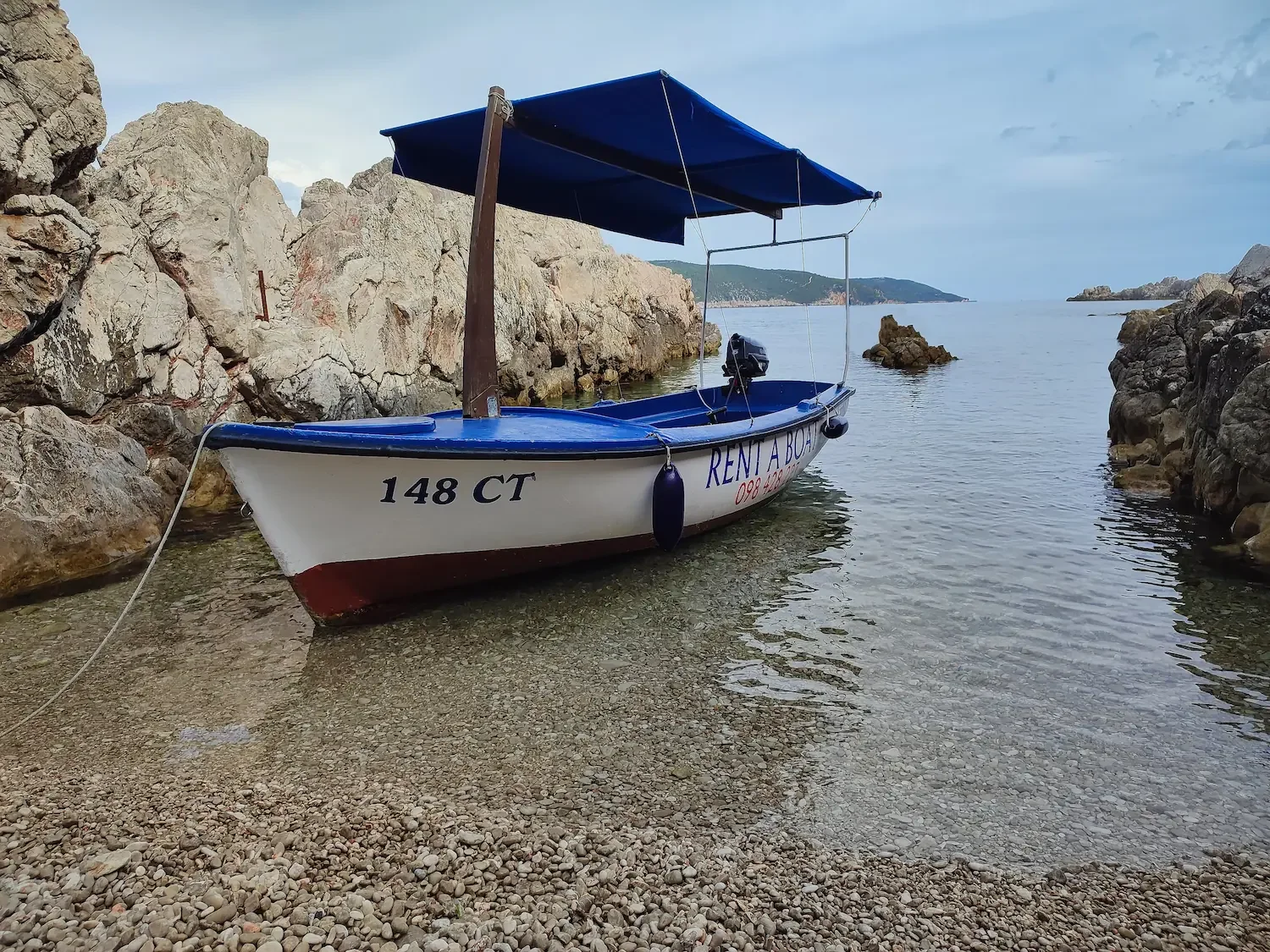 Traditional white Pasara boat 148 CT with blue sunshade parked on a pebble beach in Cavtat.
