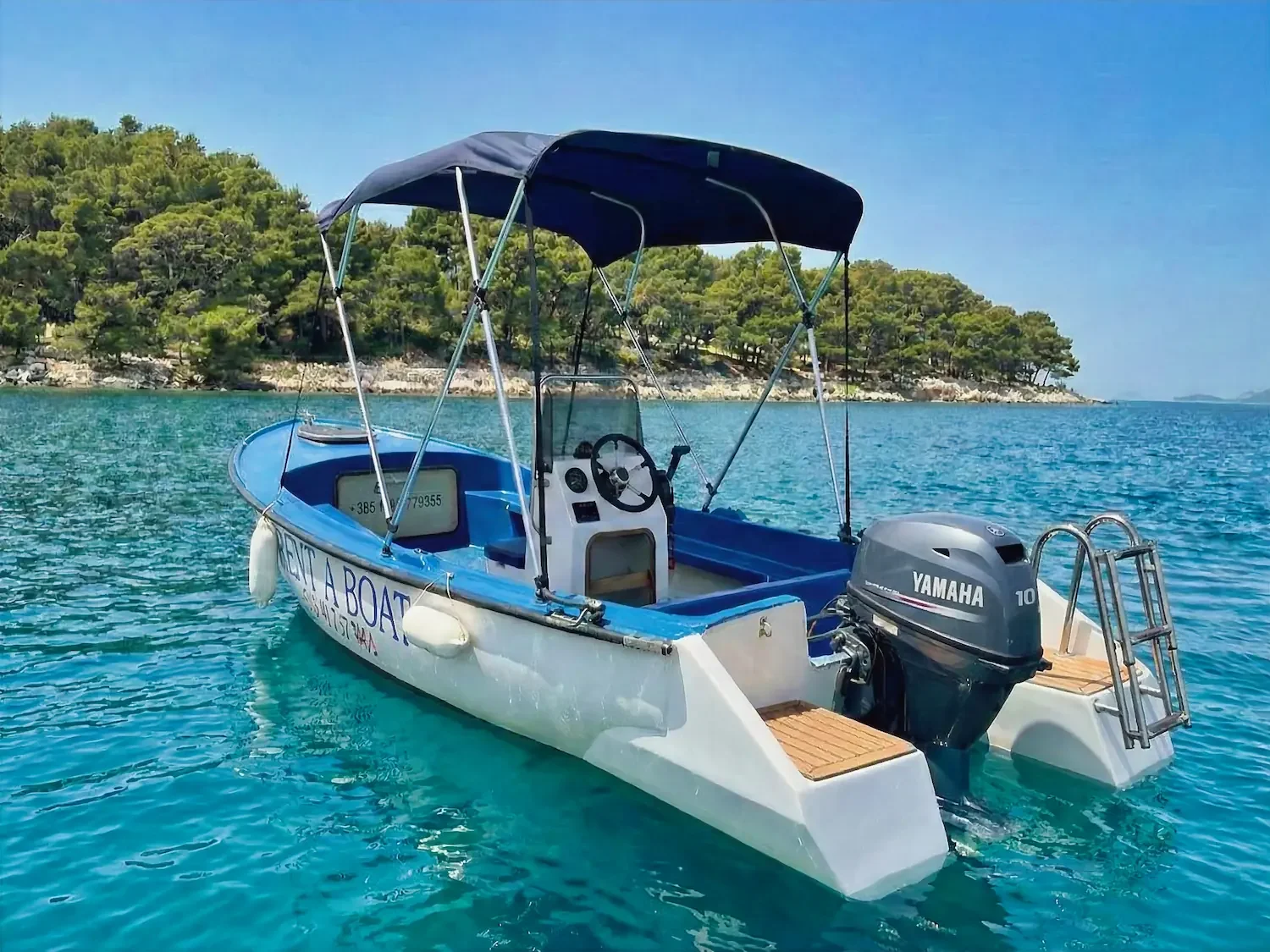 A small blue and white boat with a Yamaha outboard motor floating on calm turquoise water near a green, tree-covered shoreline under a clear blue sky.