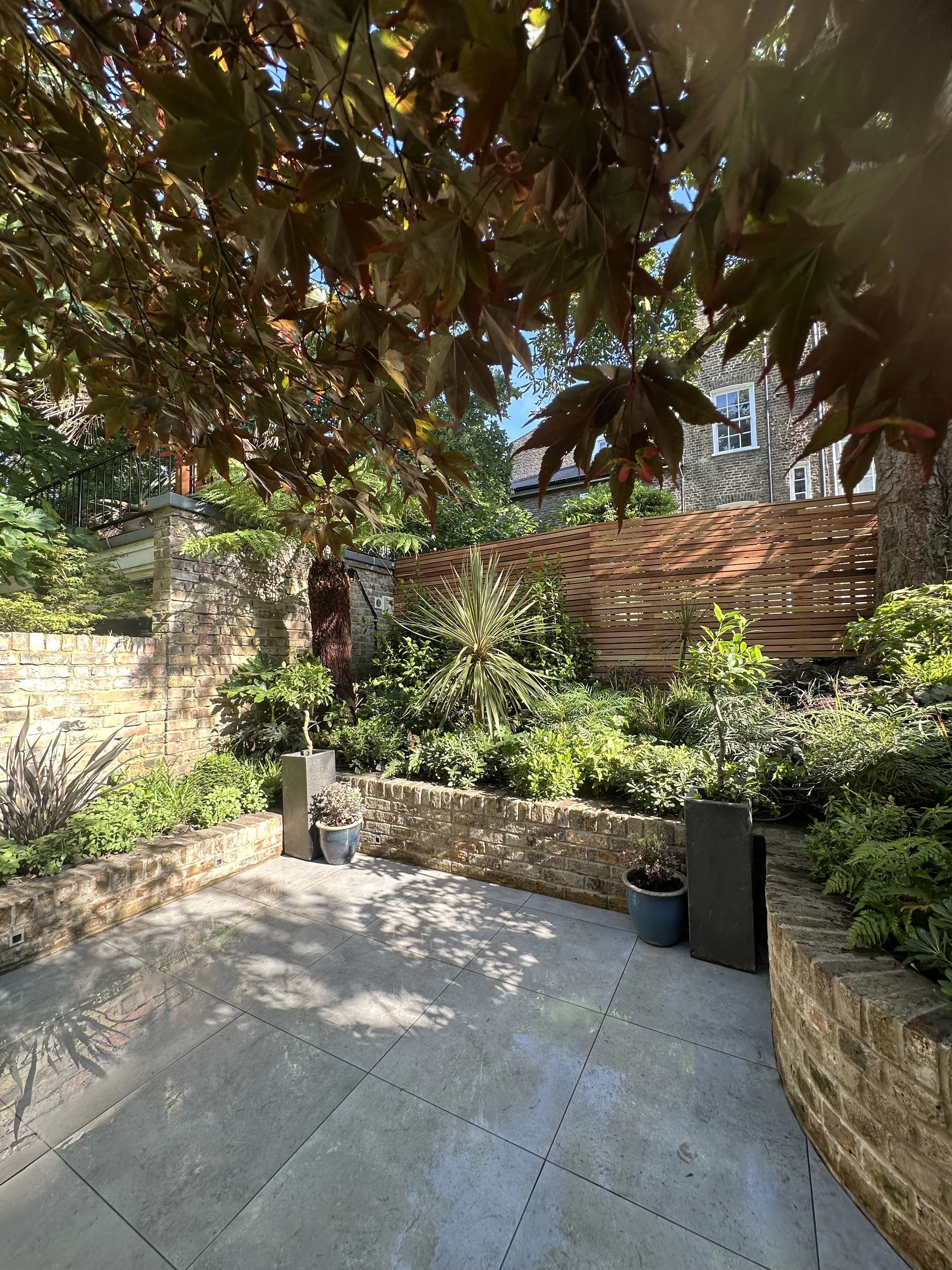 A courtyard garden with potted plants, brick walls, and a wooden fence, shaded by tree branches.
