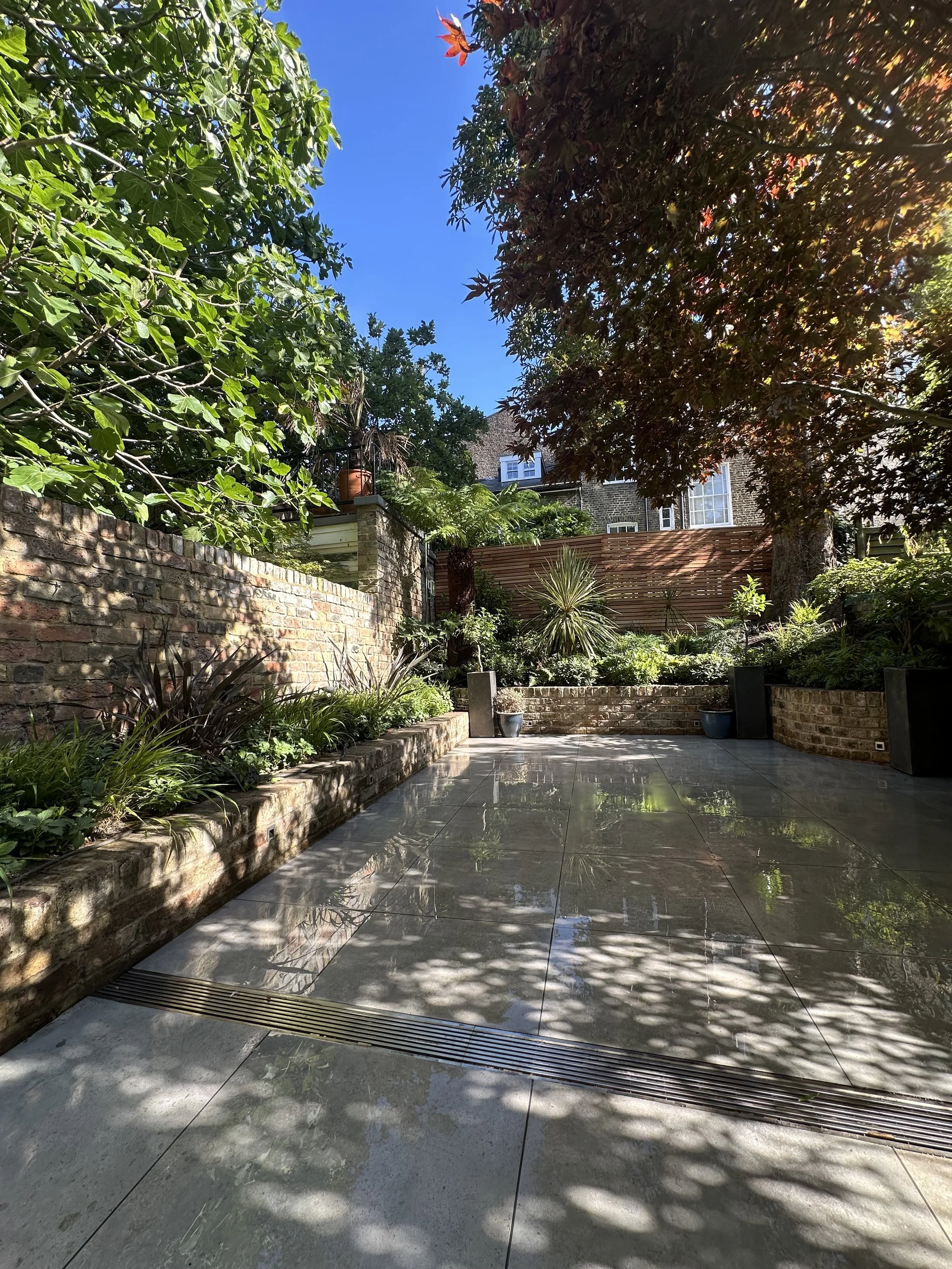 A backyard patio with tiles, surrounded by brick and stone walls with lush green plants and trees, under a clear blue sky with sunlight filtering through the leaves, creating shadows on the ground.