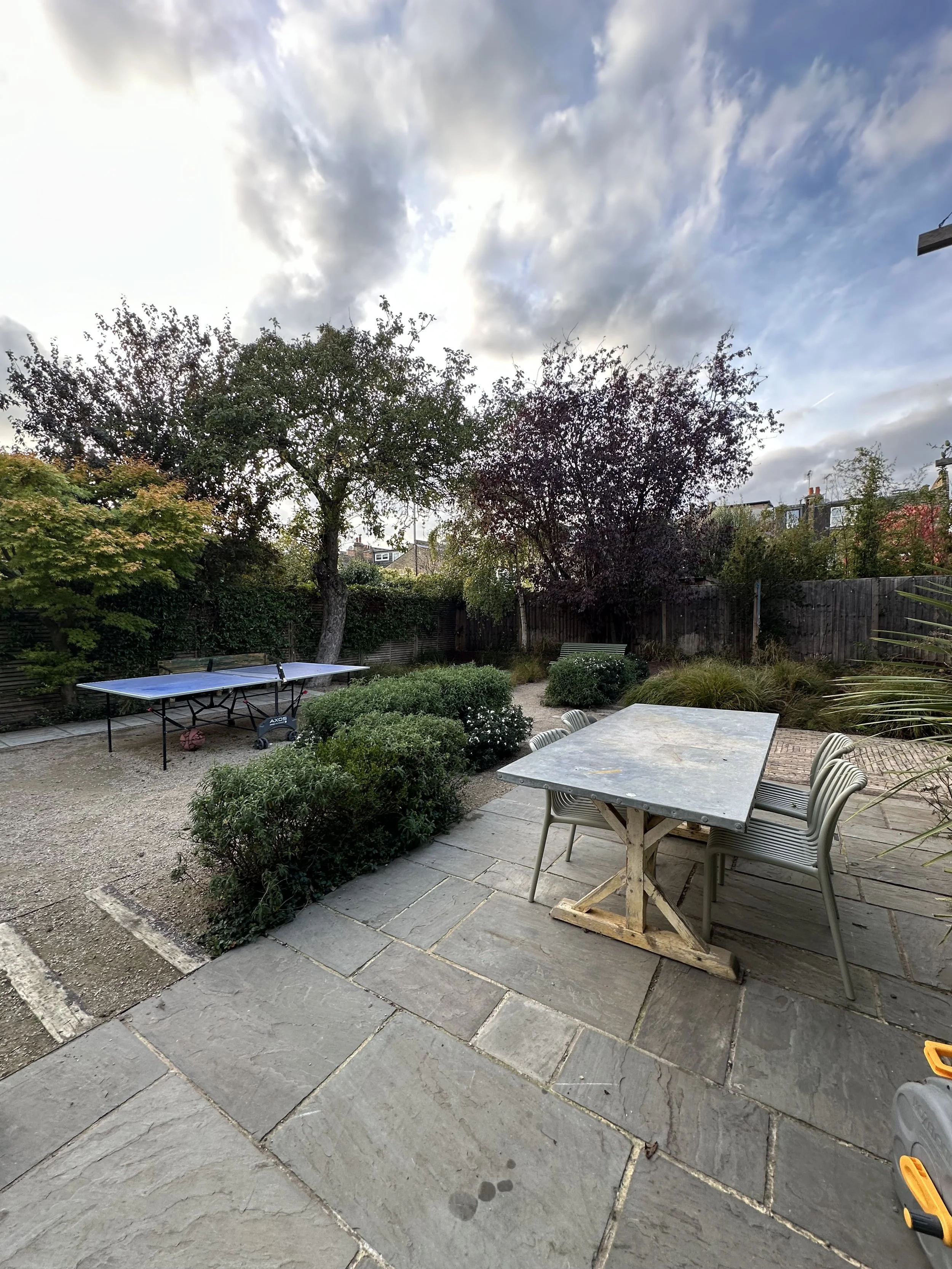 Backyard garden with a table and chairs on a stone patio, a ping pong table, trees, shrubs, and a cloudy sky.