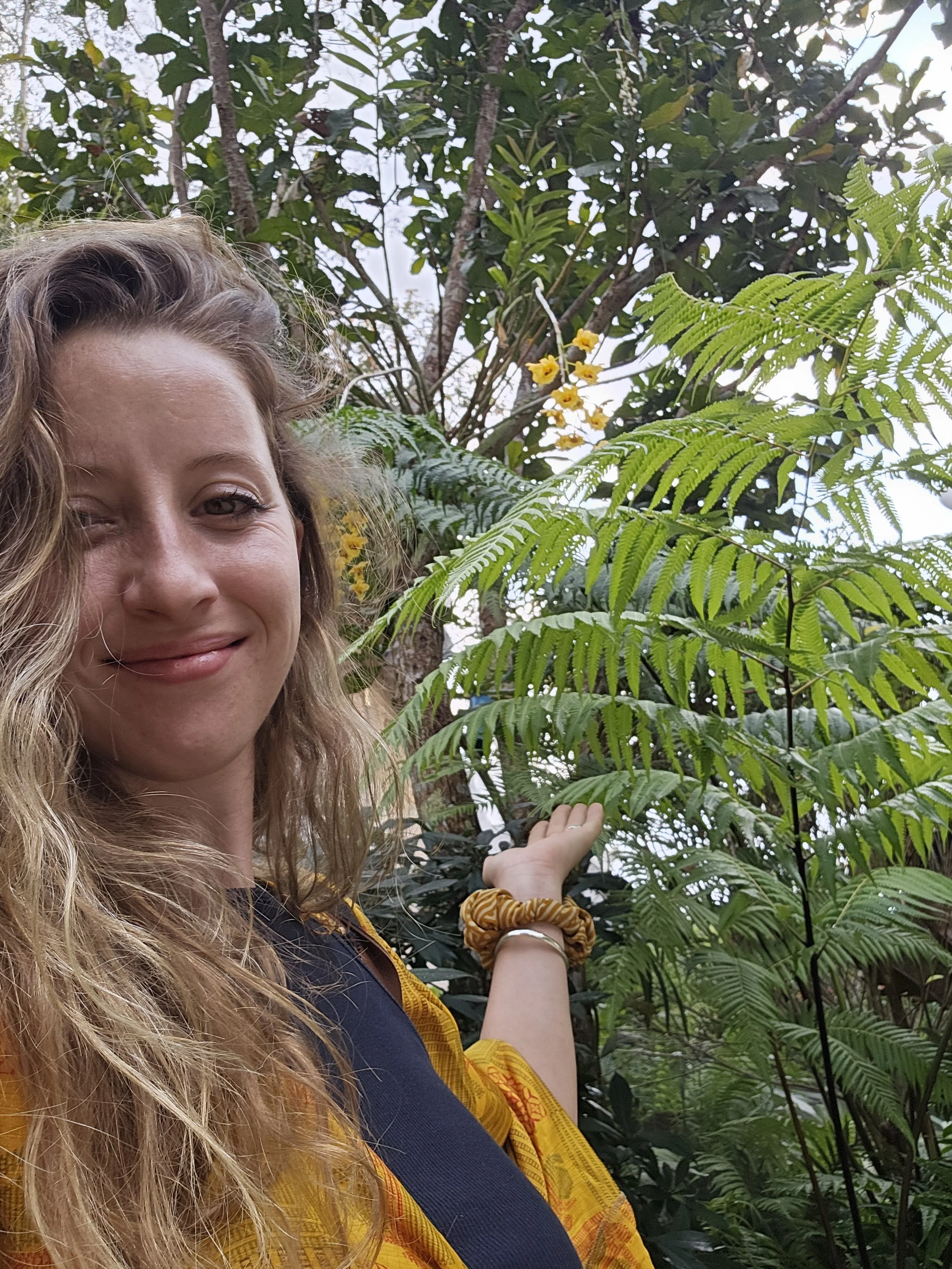 A woman with long, curly blonde hair smiling in a lush, green outdoor setting, pointing at a plant with fern-like leaves and small yellow flowers.