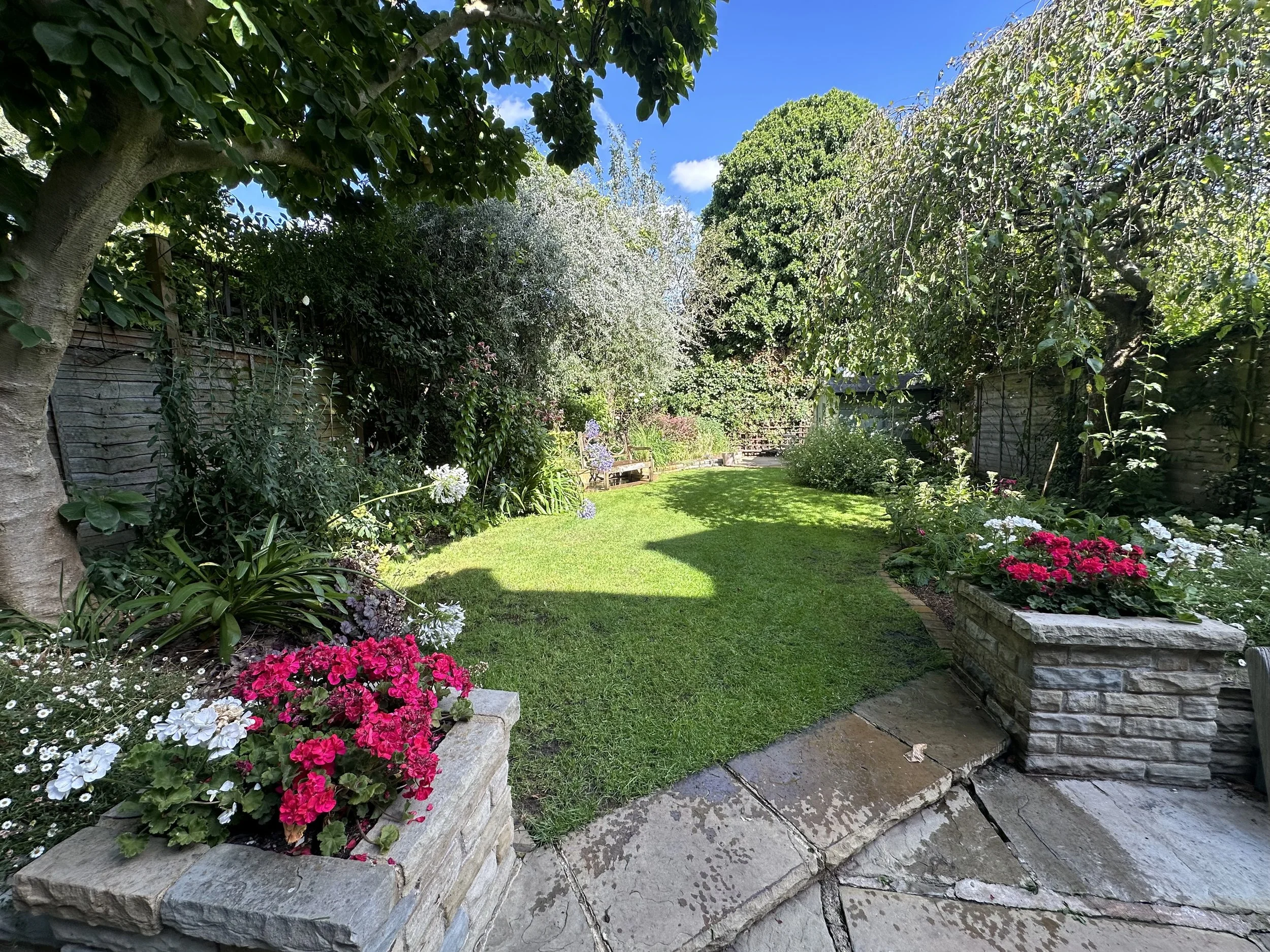 A sunny backyard garden with a well-maintained lawn, flower beds with pink, white, and purple flowers, a stone patio, and trees along the fence.