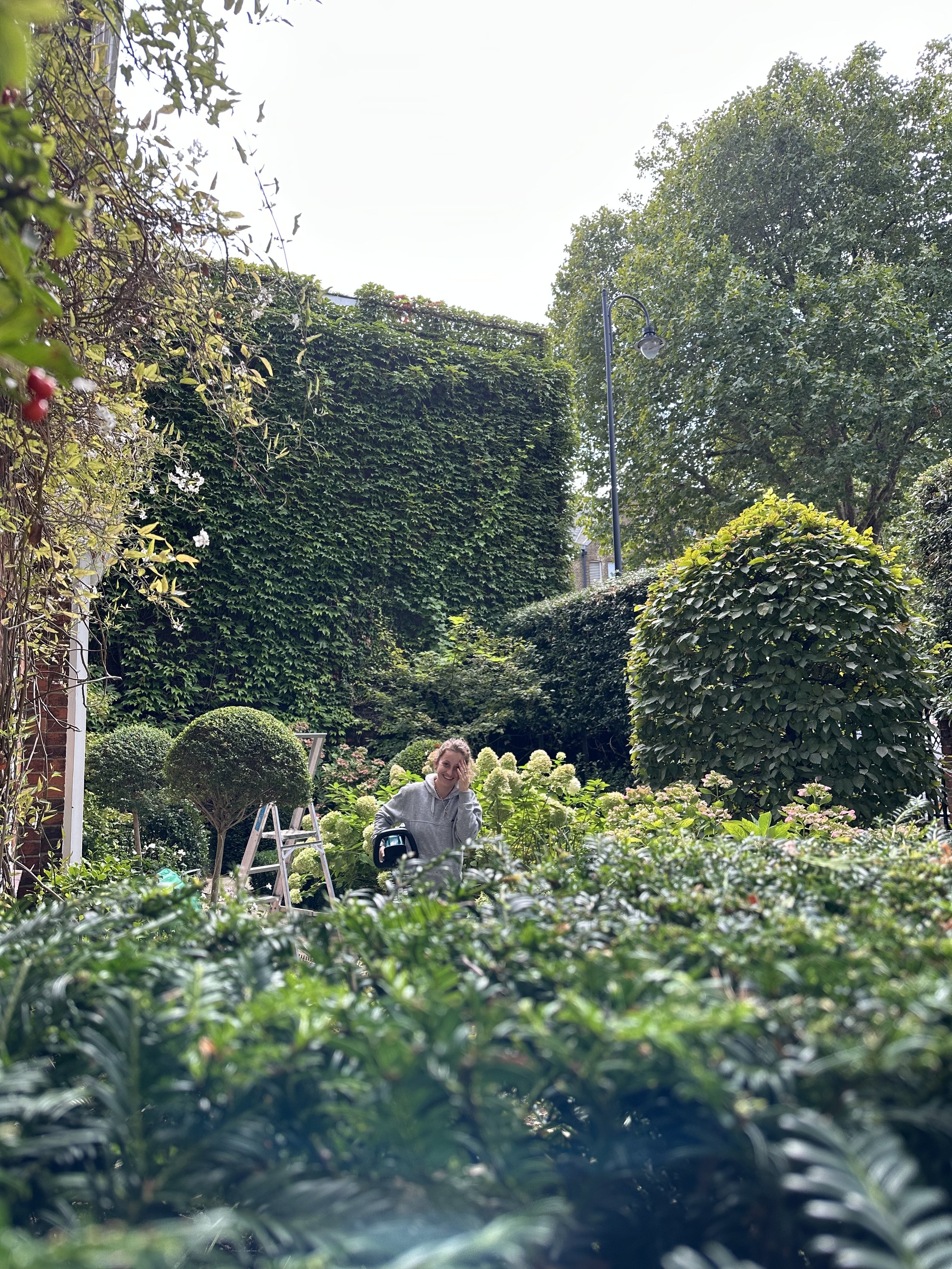Person smiling in a lush, green garden with various bushes, trees, and flowering plants.