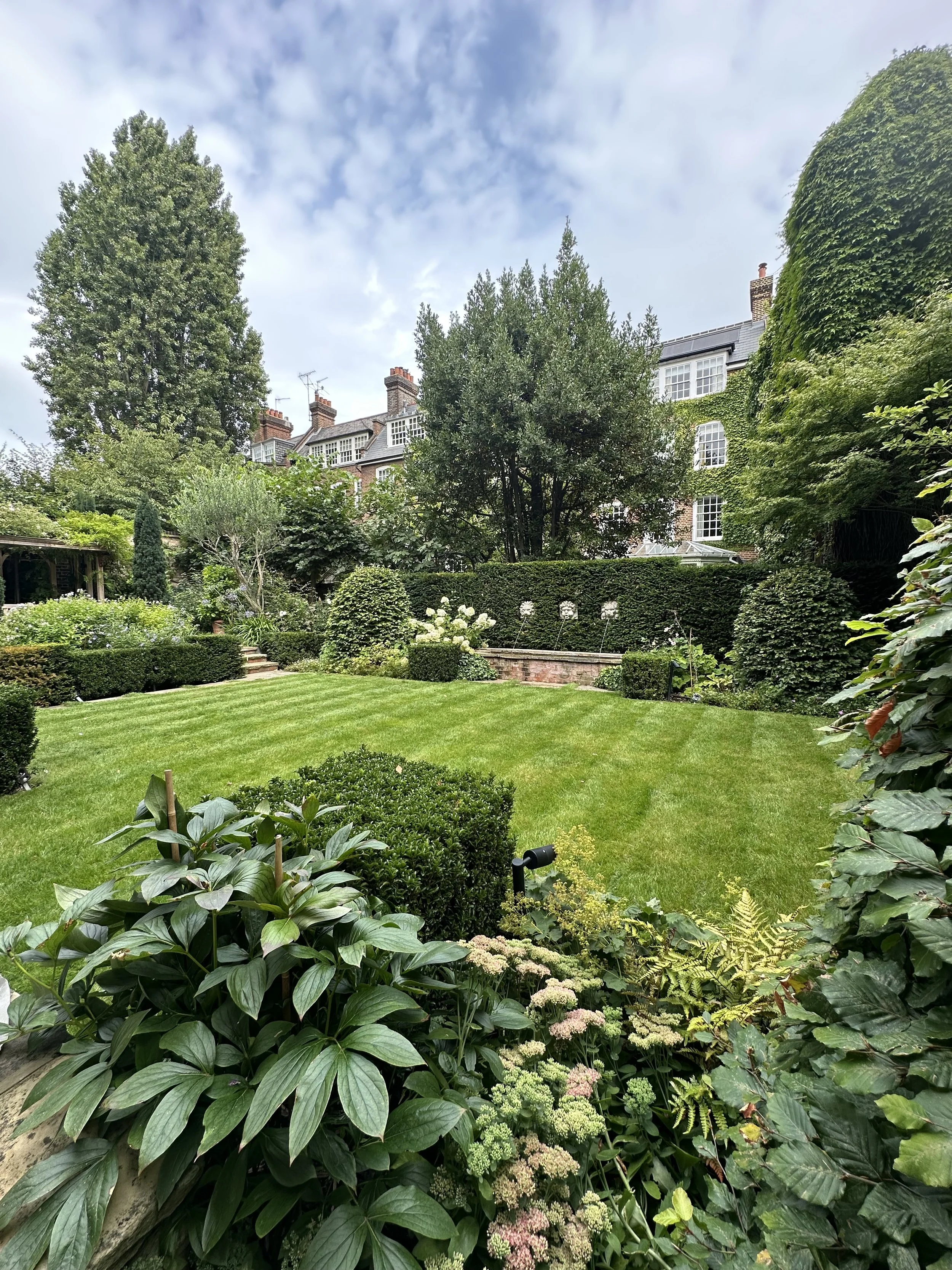 A lush, well-maintained backyard garden with green grass, various shrubs, bushes, and trees, surrounded by a tall brick house with numerous windows, and a partly cloudy sky overhead.