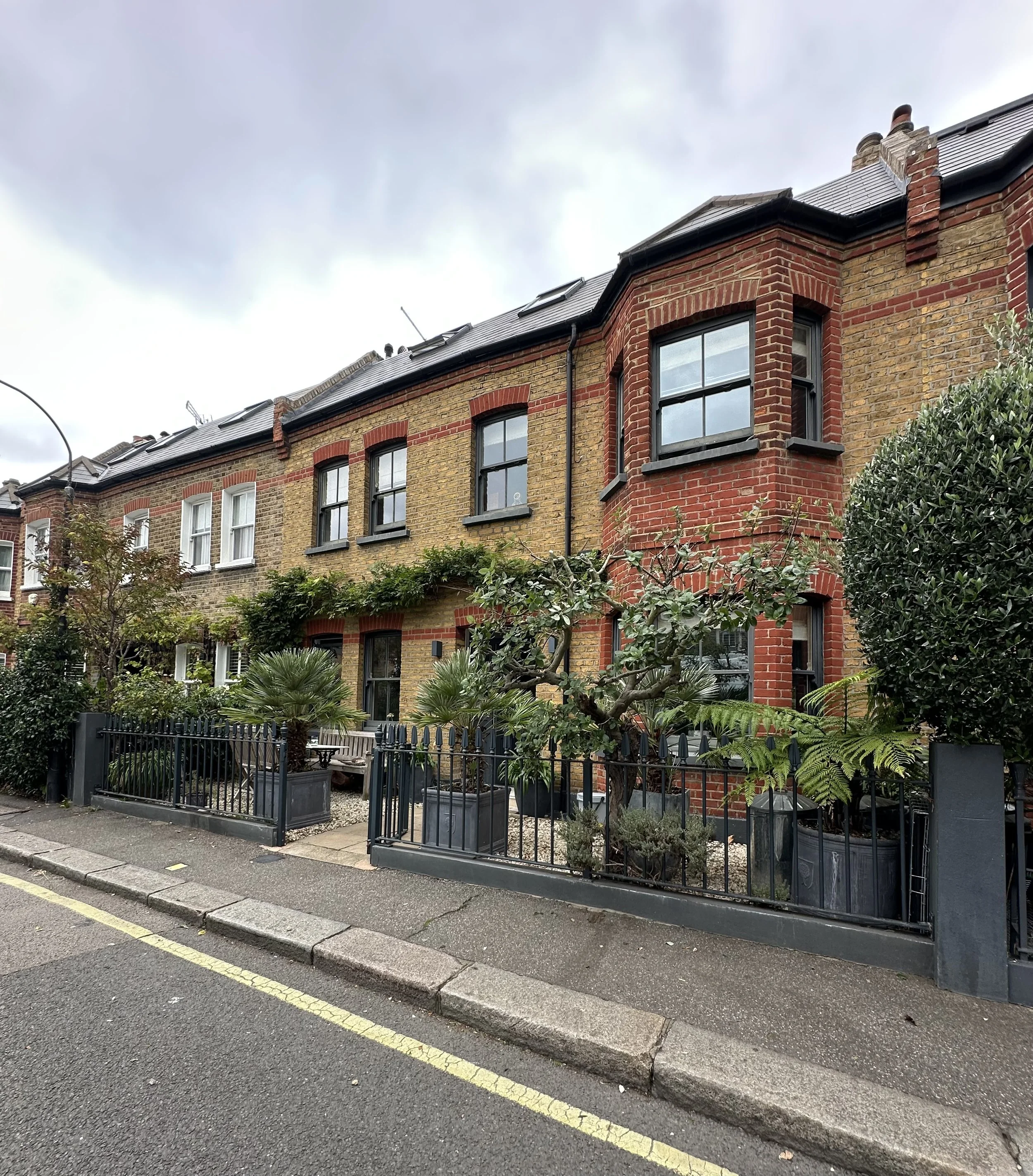 A row of brick townhouses with black-framed windows, surrounded by potted plants and greenery, located along a paved sidewalk under an overcast sky.