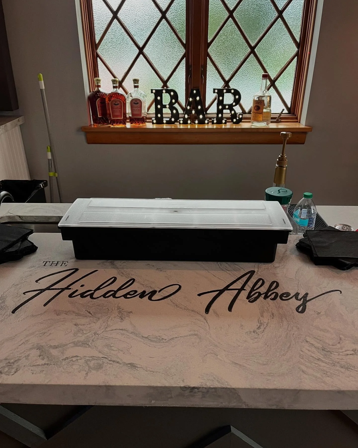 Bar area with bottles of whiskey, decorative 'BAR' sign, behind a window, and a counter with a sink and bottled water, with 'The Hidden Abbey' written on the counter.