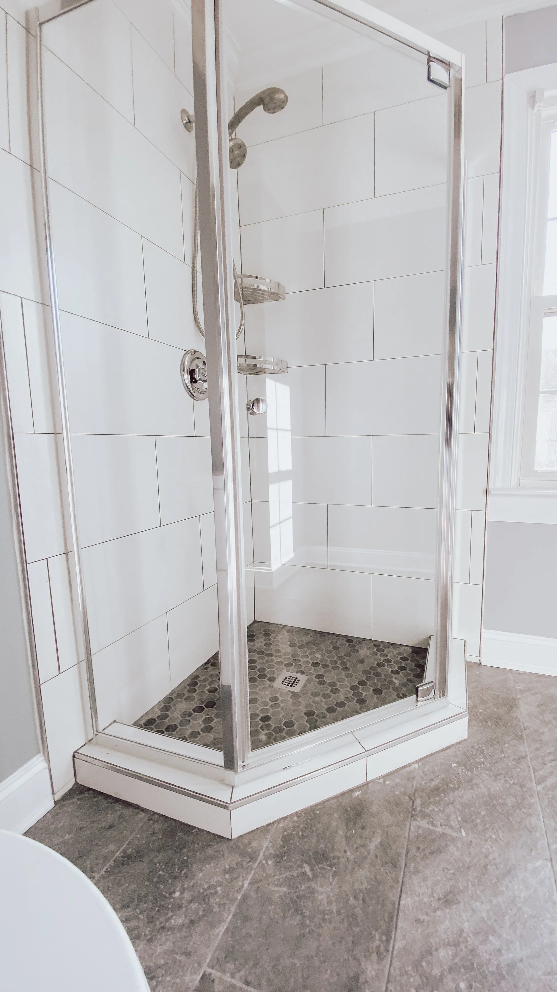 A small enclosed shower with clear glass door, chrome fixtures, white tiled walls, and a dark hexagonal tiled shower floor, next to a window and a corner of a gray tiled bathroom floor.