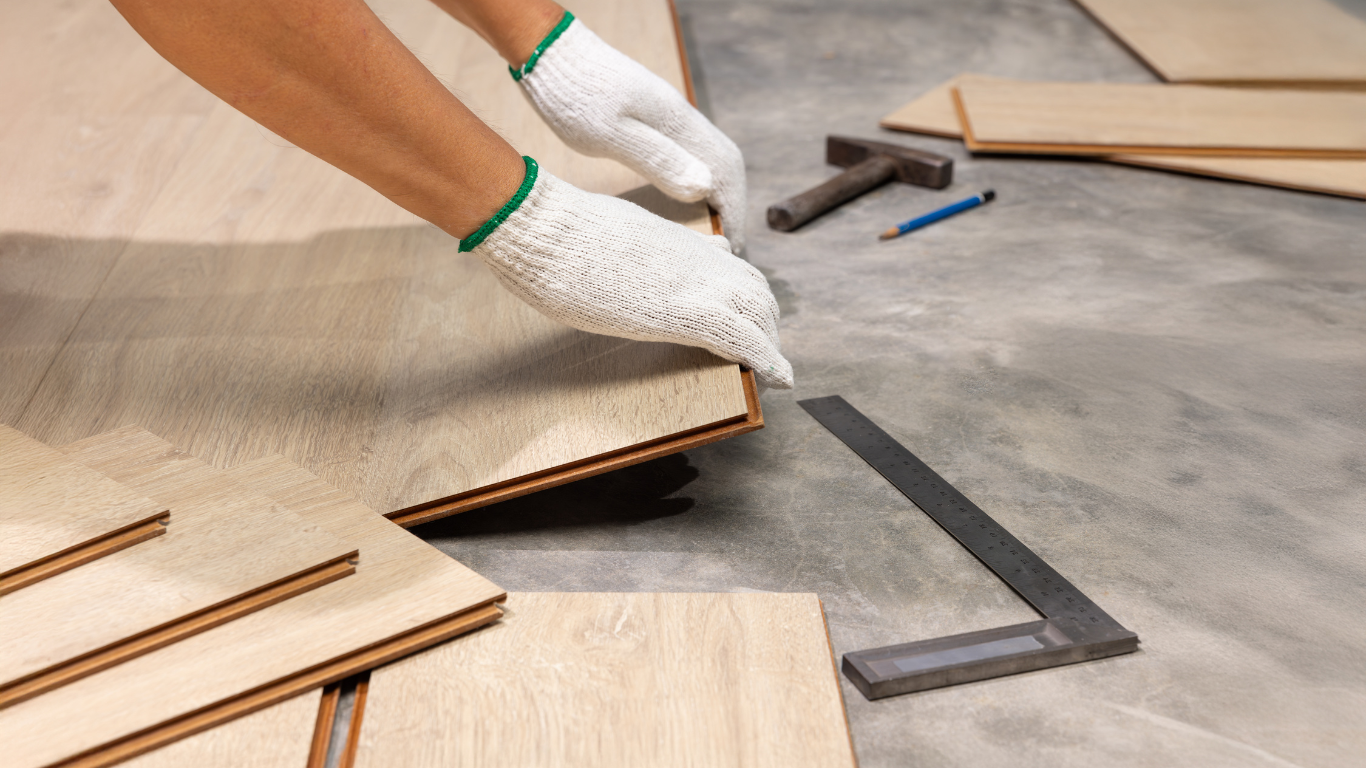 Person wearing white gloves installing wood flooring with measuring tools and wooden planks nearby.