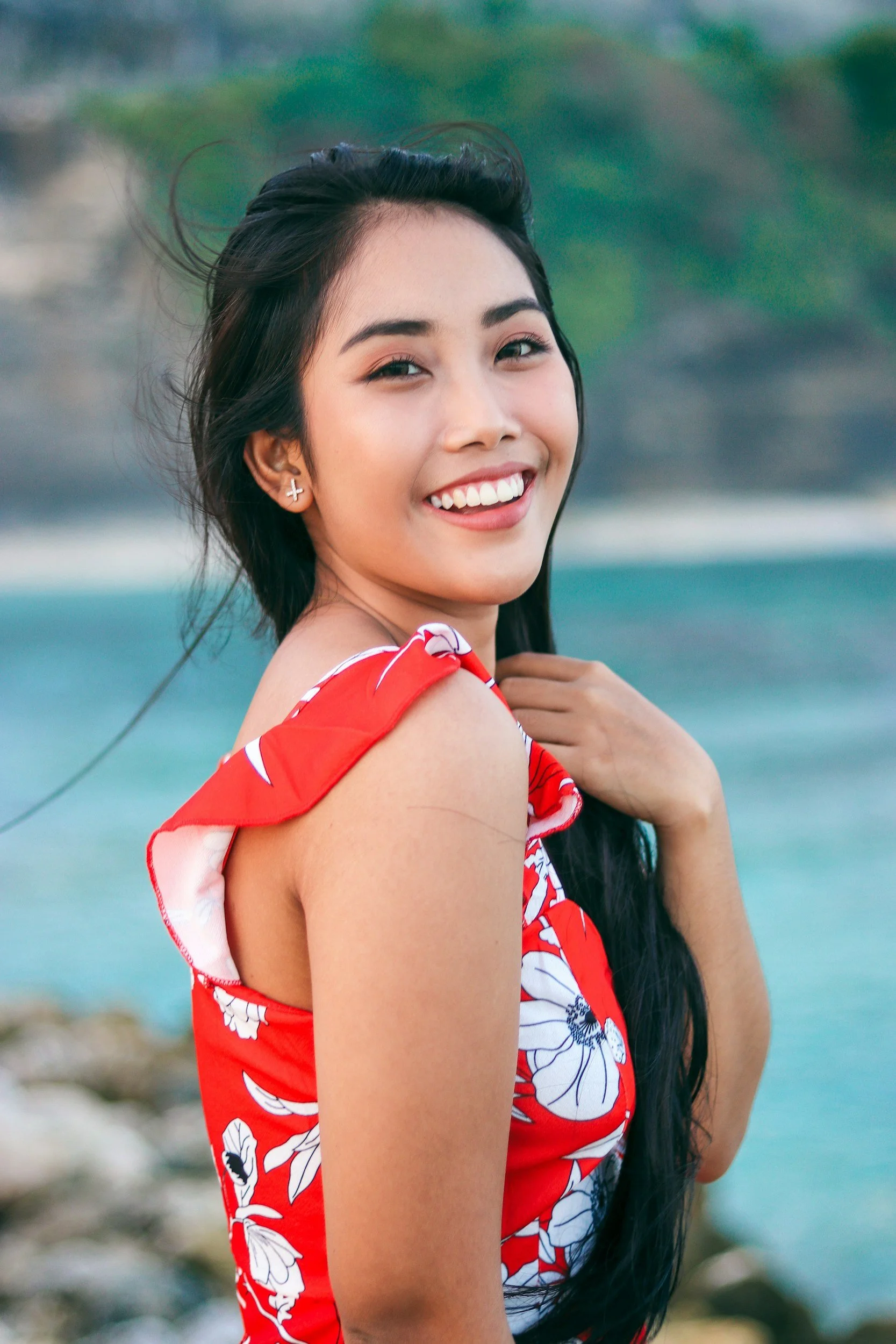 A smiling young woman with long dark hair, wearing earrings shaped like crosses, dressed in a red, white, and blue floral dress, standing outdoors near water with blurred green foliage in the background.