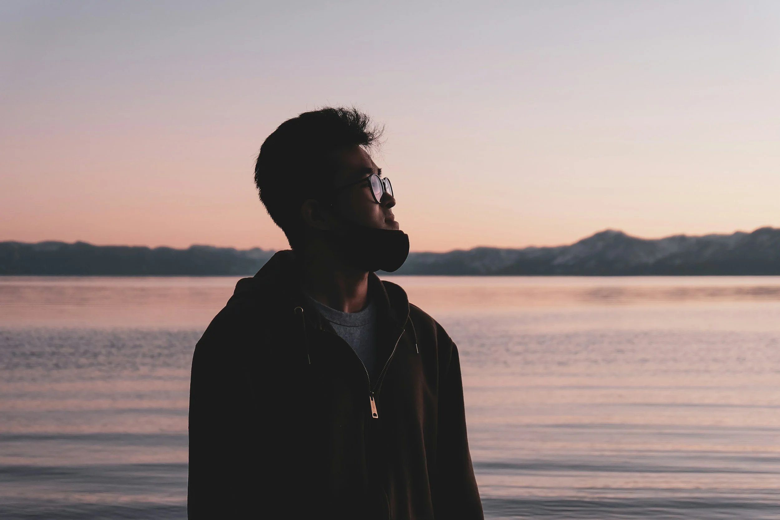 A person wearing glasses and a jacket, standing by a body of water with mountains in the background during sunset or sunrise.