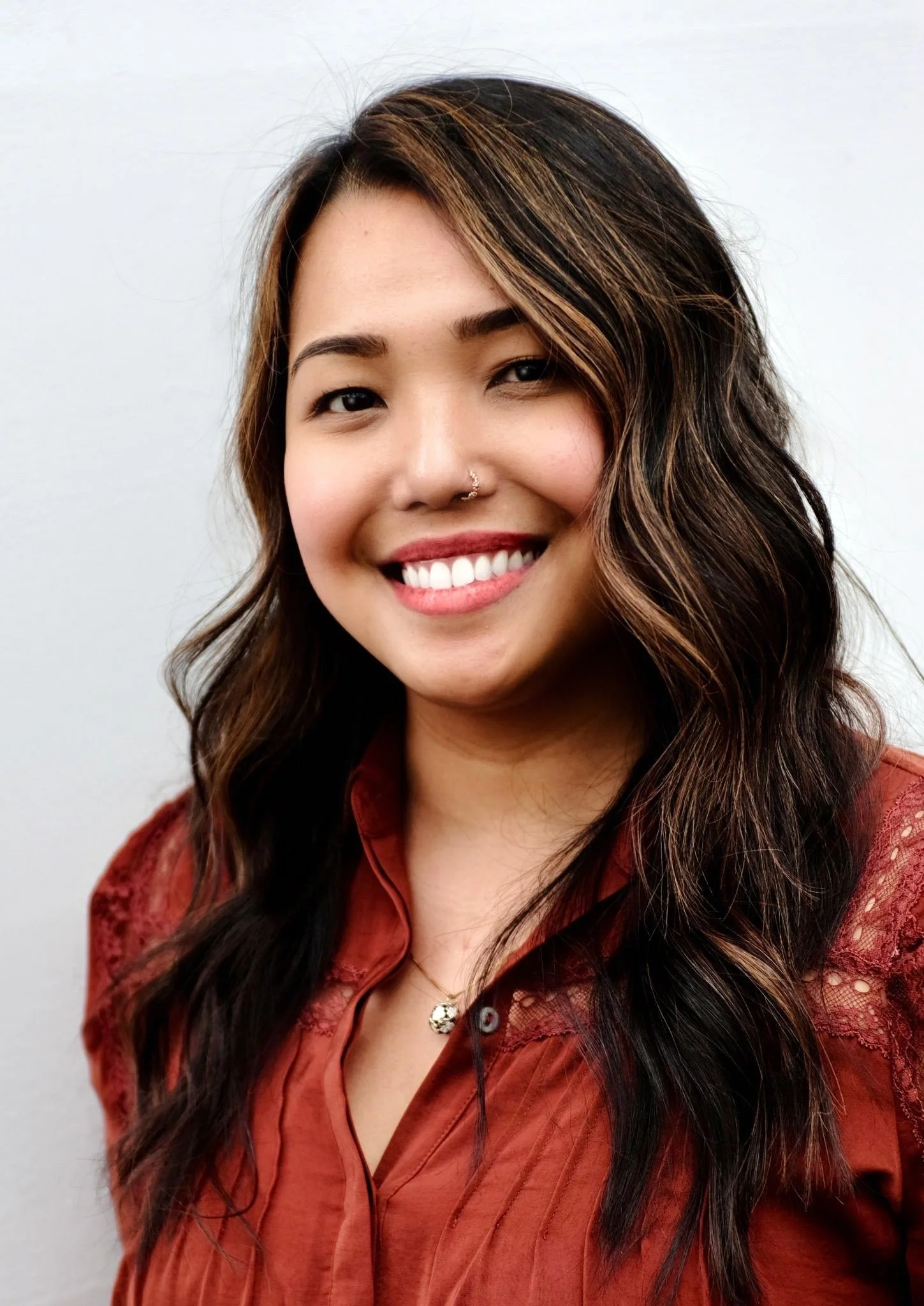 A woman with long wavy brown hair with blonde highlights, smiling, wearing a rust-colored blouse with lace details, standing against a plain light-colored background.