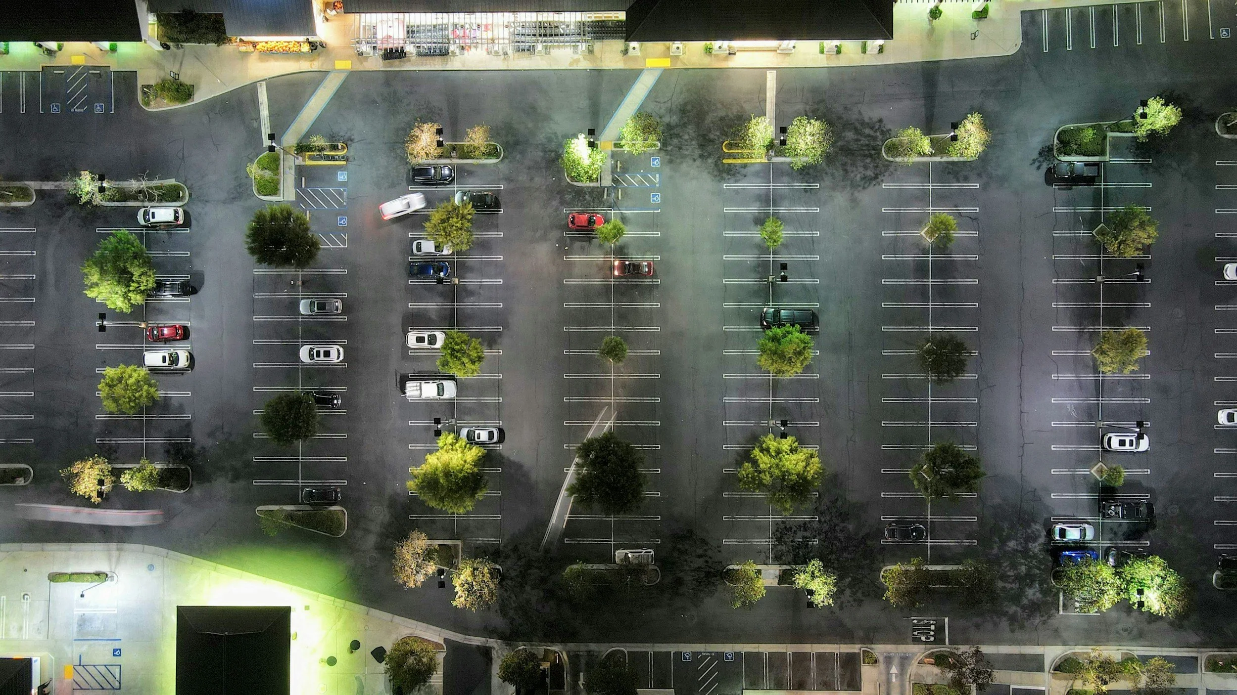 An aerial view of an empty parking lot at night with scattered cars, trees and illuminated storefronts.