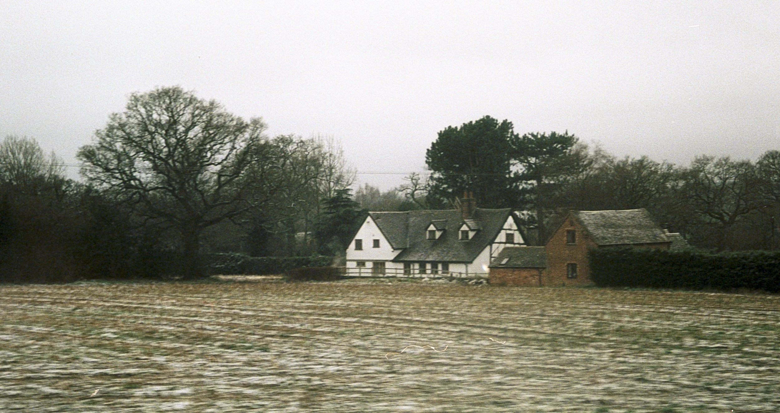 A rural landscape with flood waters encompassing the field and several houses, under an overcast sky.