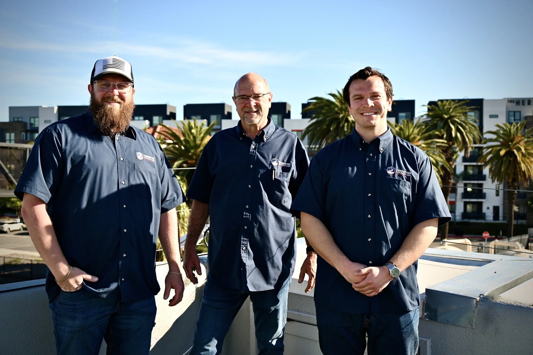 Three men wearing blue shirts standing outdoors on a rooftop with palm trees and buildings in the background.
